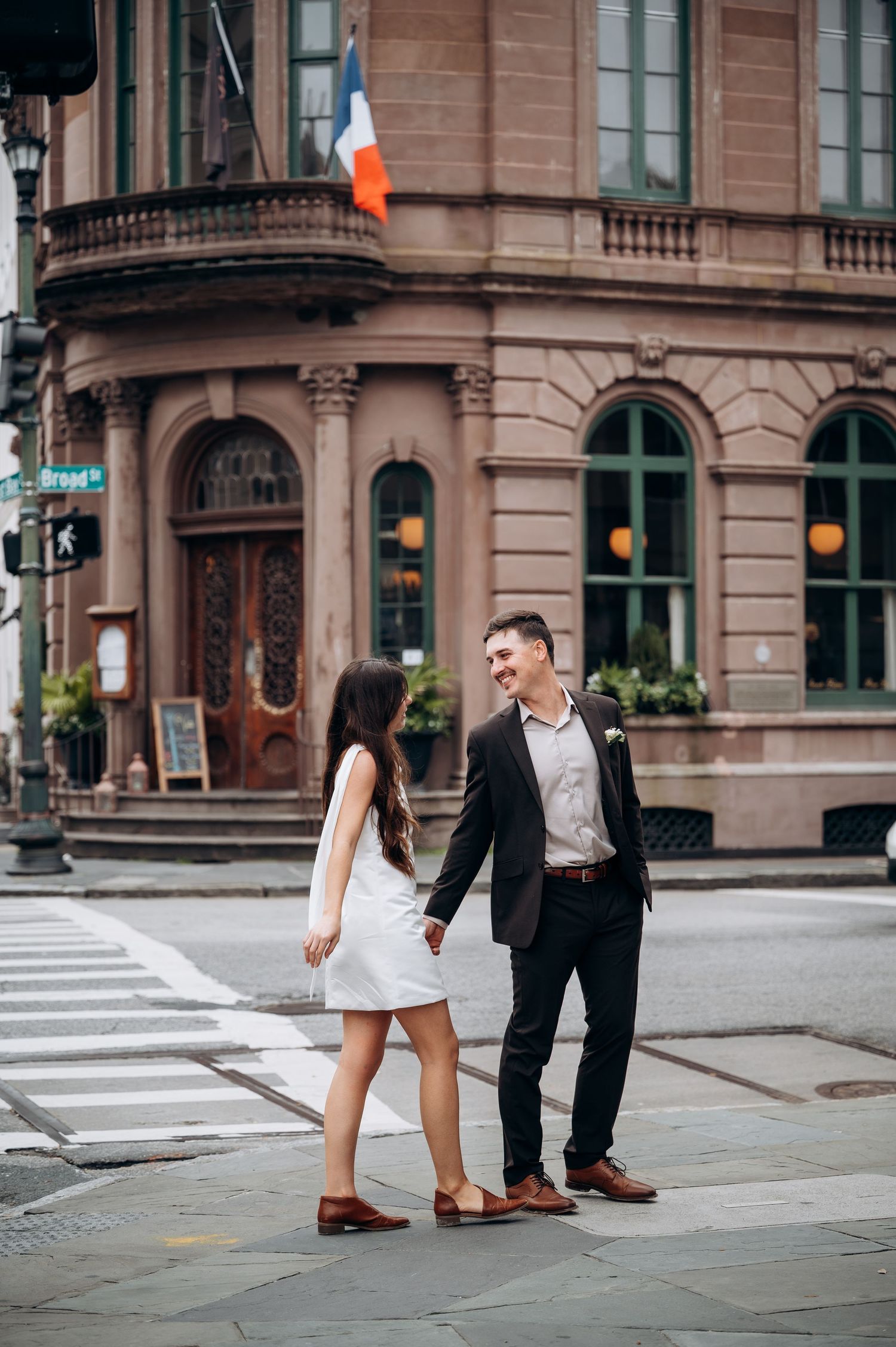 A couple poses playfully at an urban street corner with historic buildings in the background.