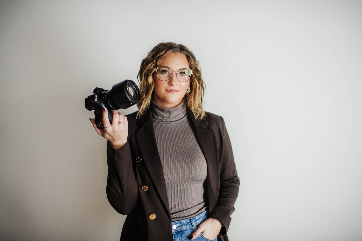 Photographer in brown blazer poses with camera against white background.