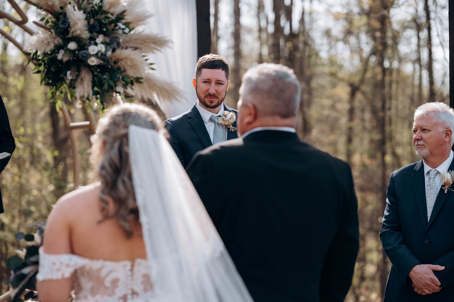 Outdoor wedding ceremony setup with white draping and floral arrangements in wooded setting.