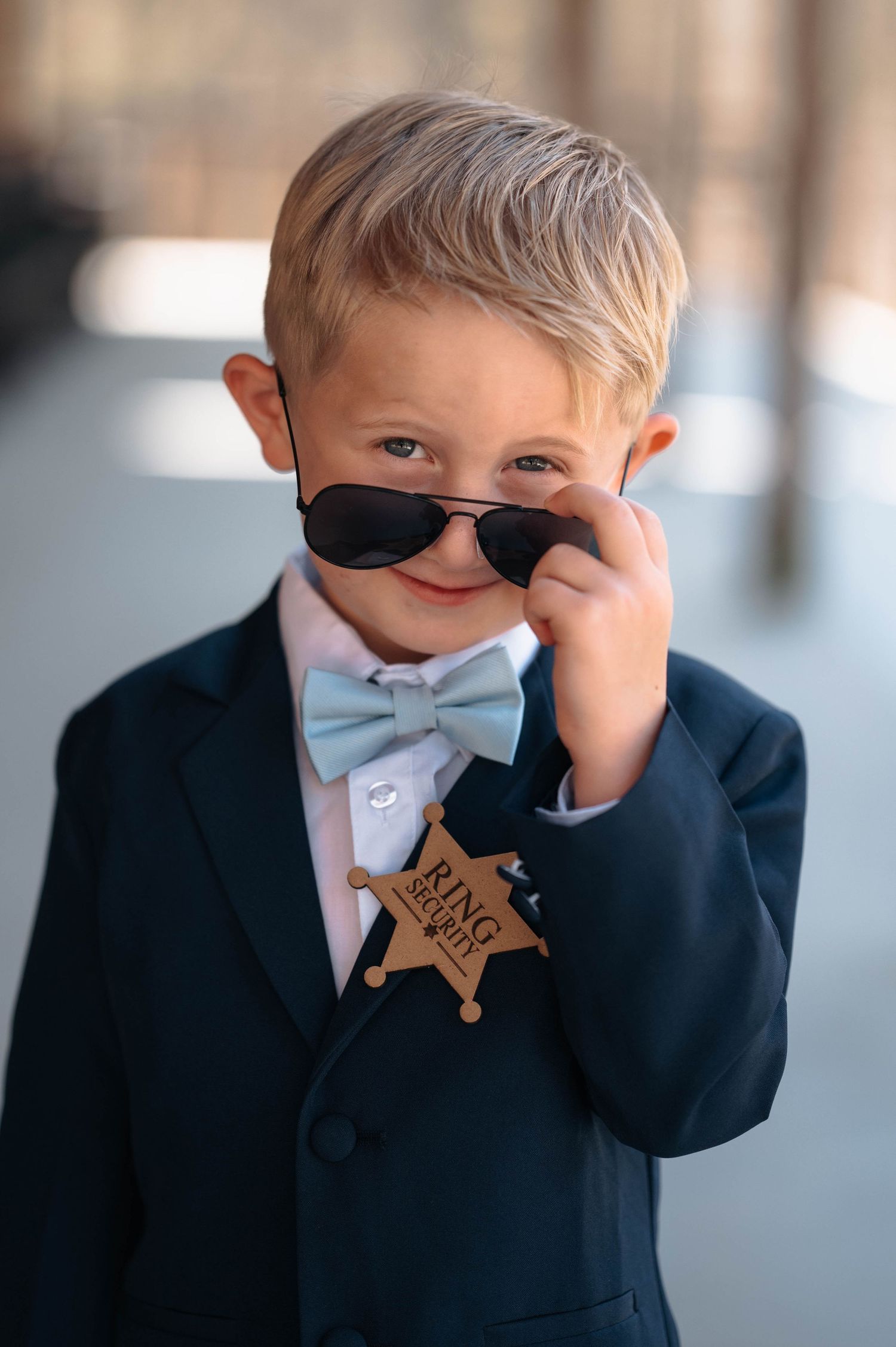 Young ring bearer in navy suit poses playfully with sunglasses and security badge.