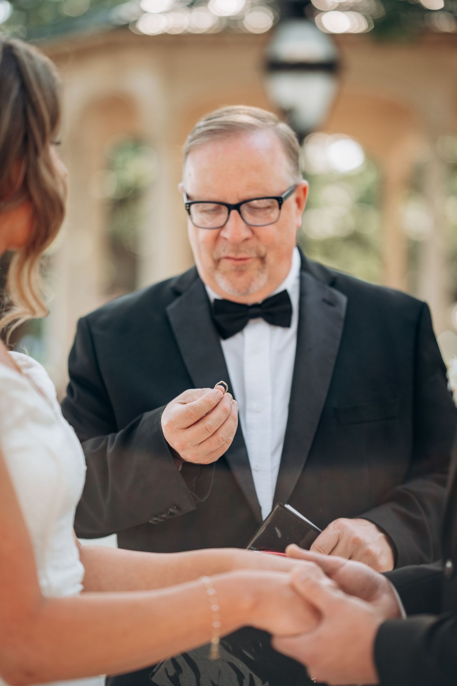 Officiant in black tuxedo holding wedding ring during outdoor ceremony ring exchange.