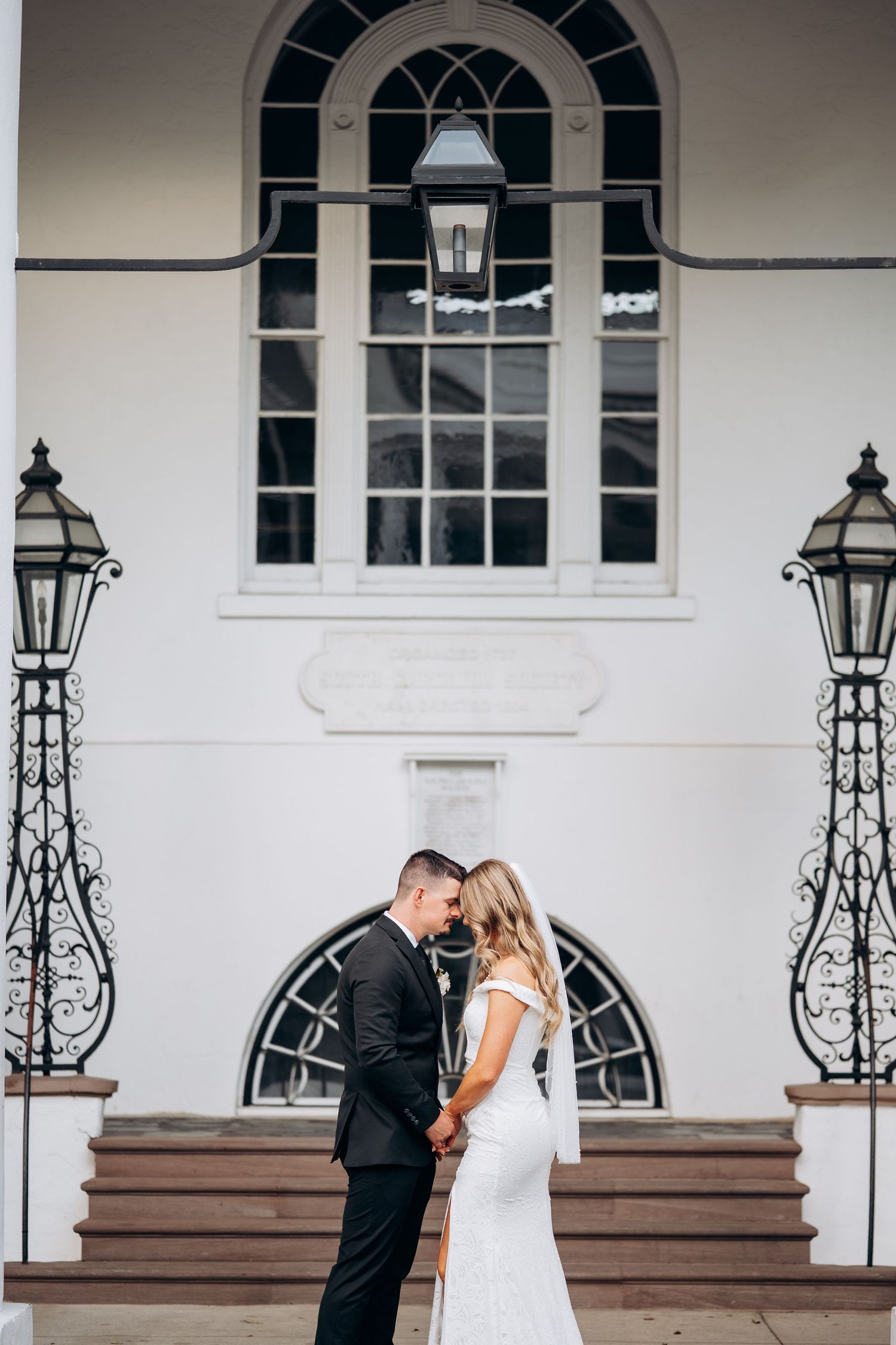 Bride and groom share intimate moment holding hands outside white historic wedding venue.
