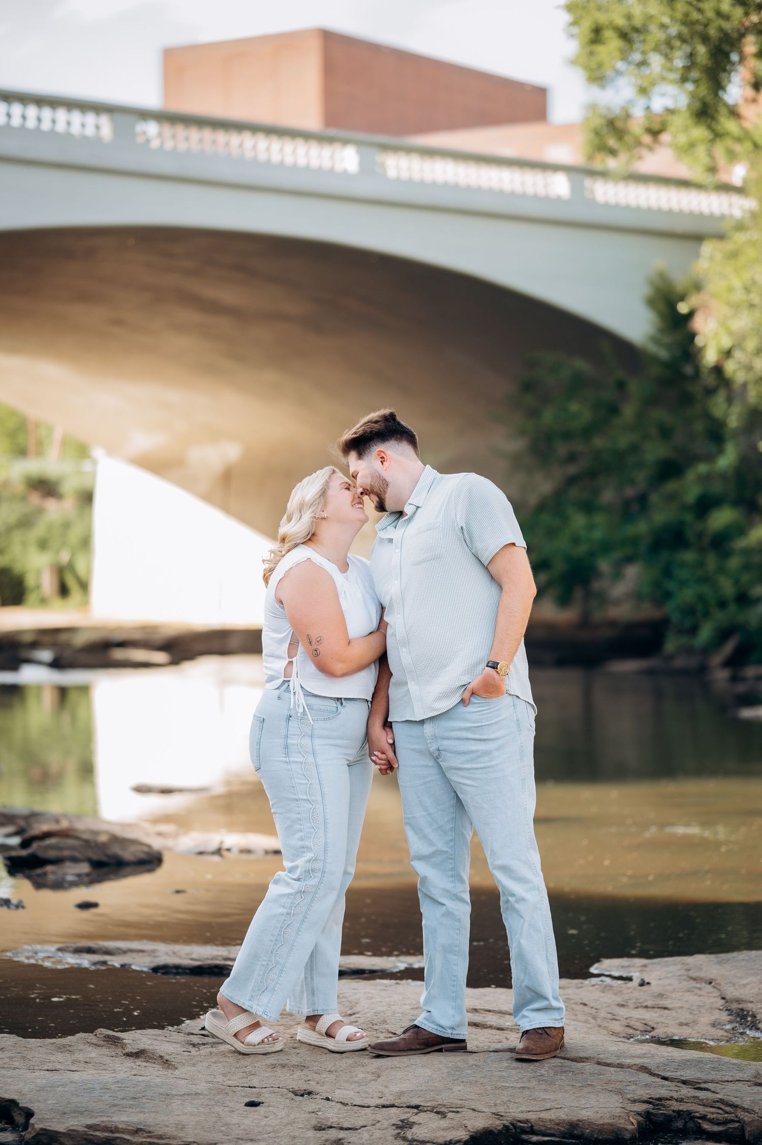 Couple sharing a romantic kiss by a river with a bridge backdrop during golden hour.