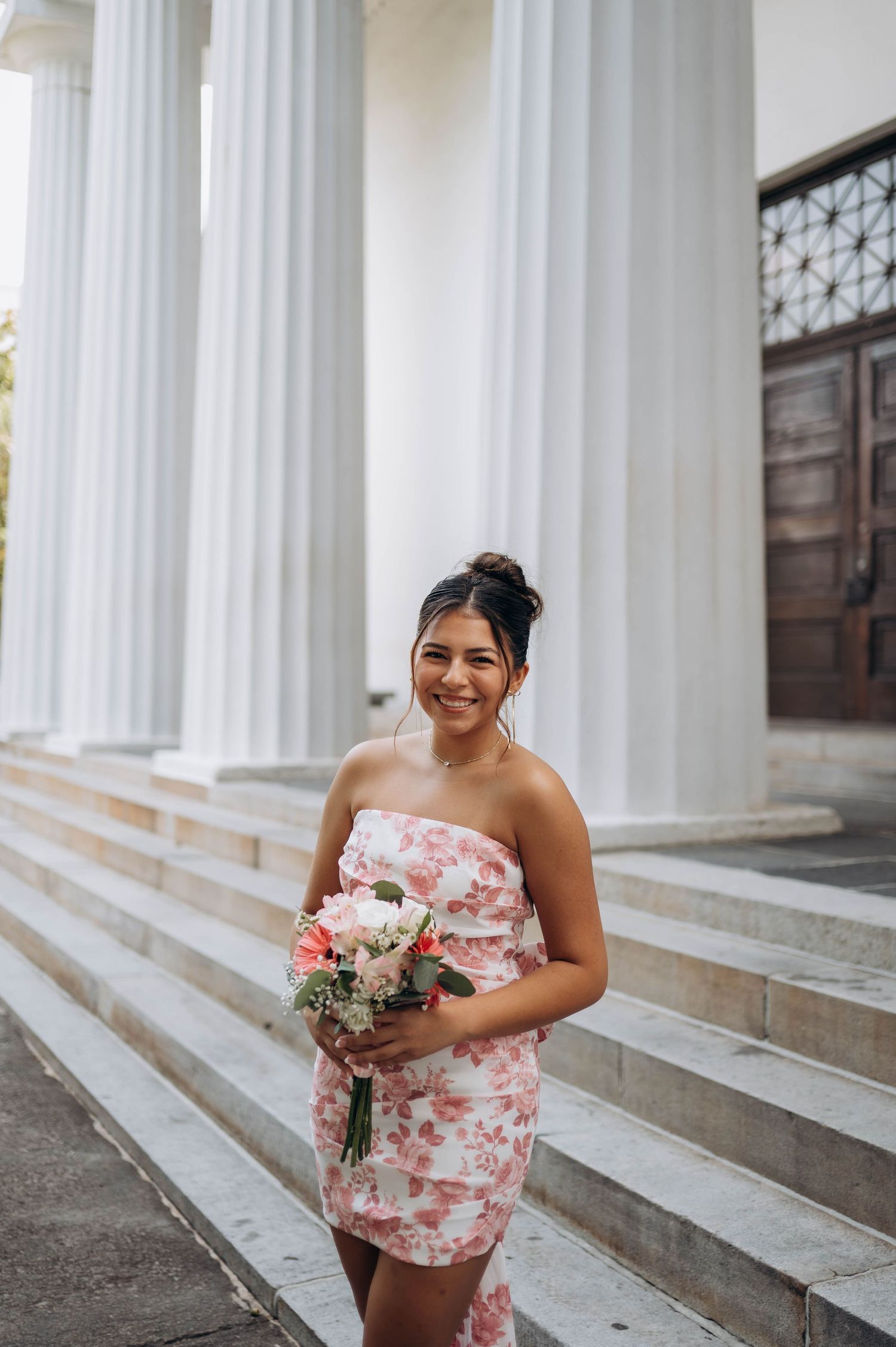 A person in a floral dress smiling while holding a bouquet on white columned steps.