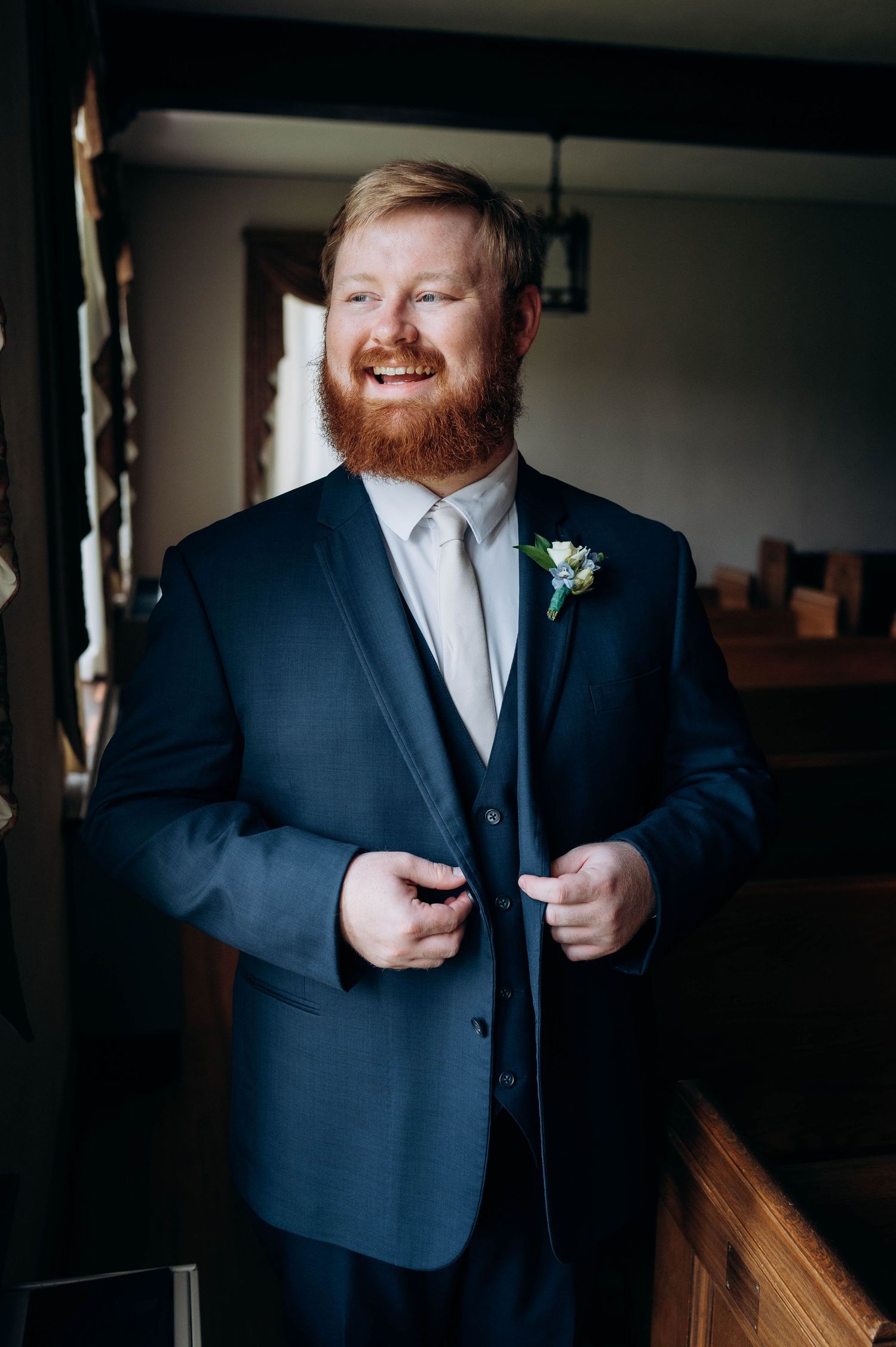 A joyful groom with red beard wearing a navy suit smiles while preparing for the ceremony.