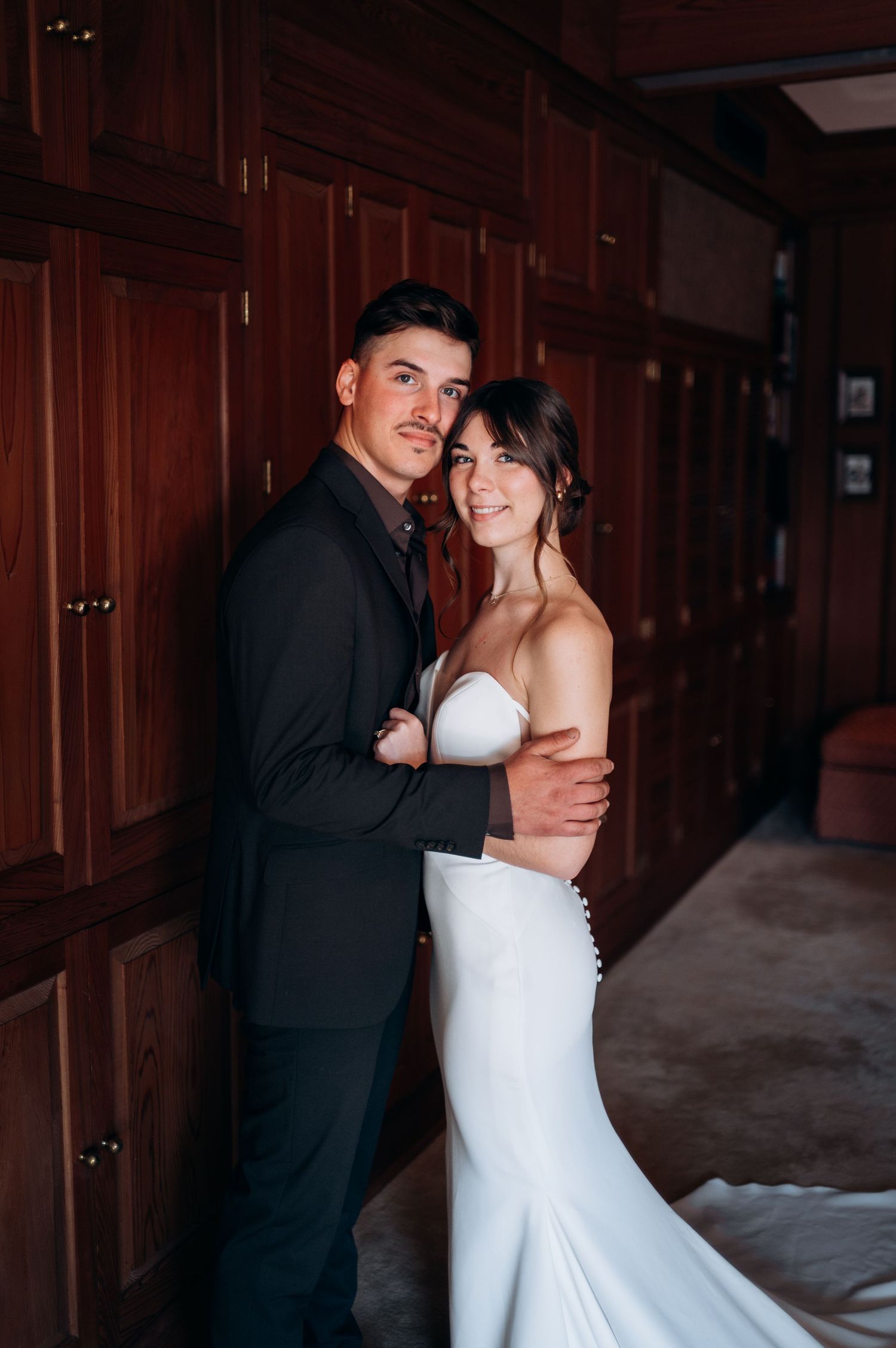 Smiling bride and groom embrace in a warmly lit wood-paneled room on their wedding day.