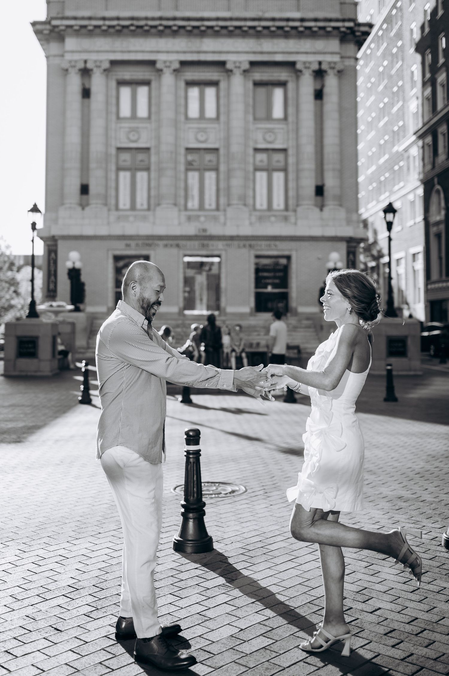 Black and white photo of a couple dancing joyfully on a city street, woman in white dress lifting her leg, urban building backdrop.