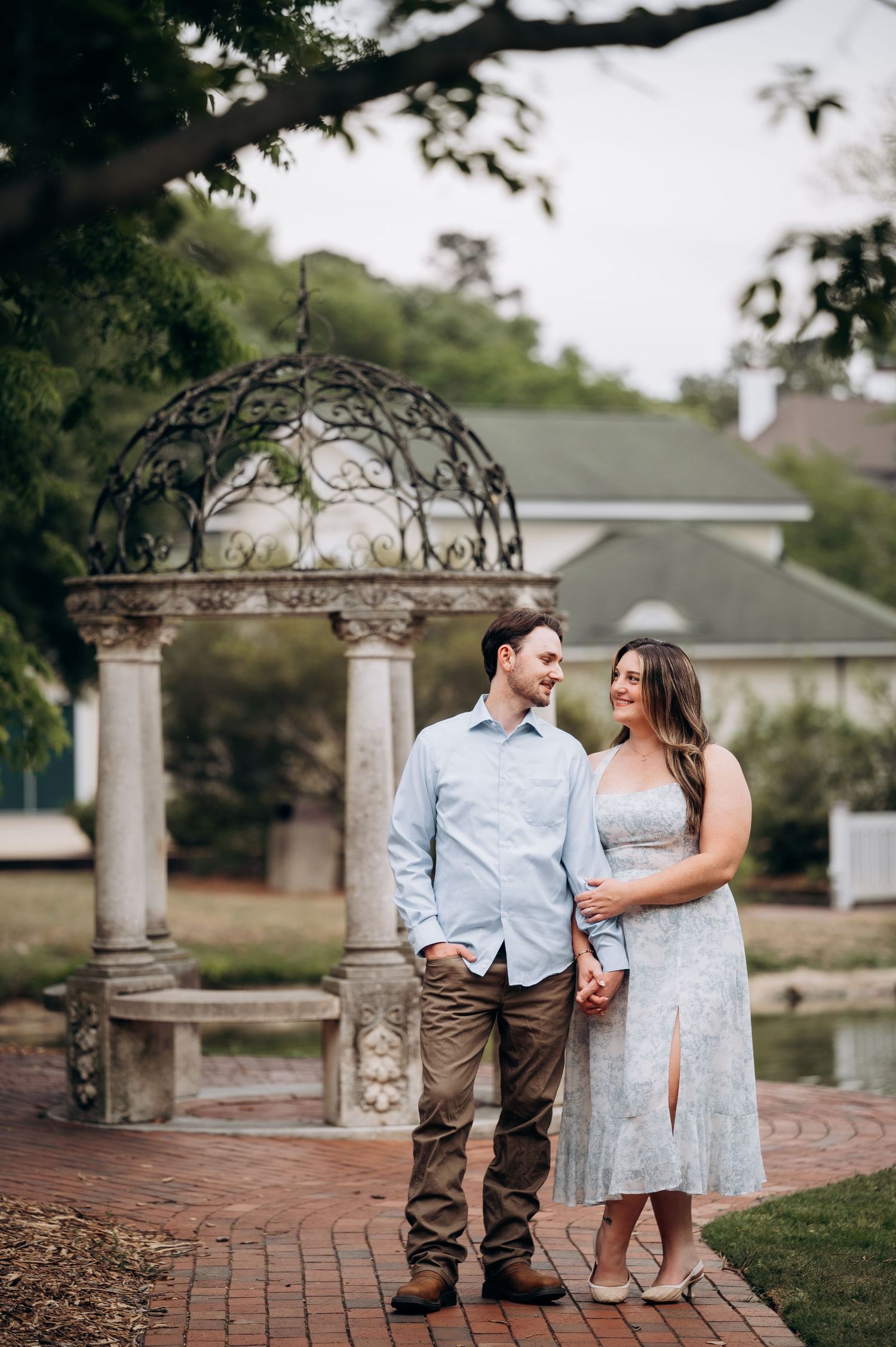 Smiling couple holding hands near an ornate stone gazebo beside a pond in a scenic garden.