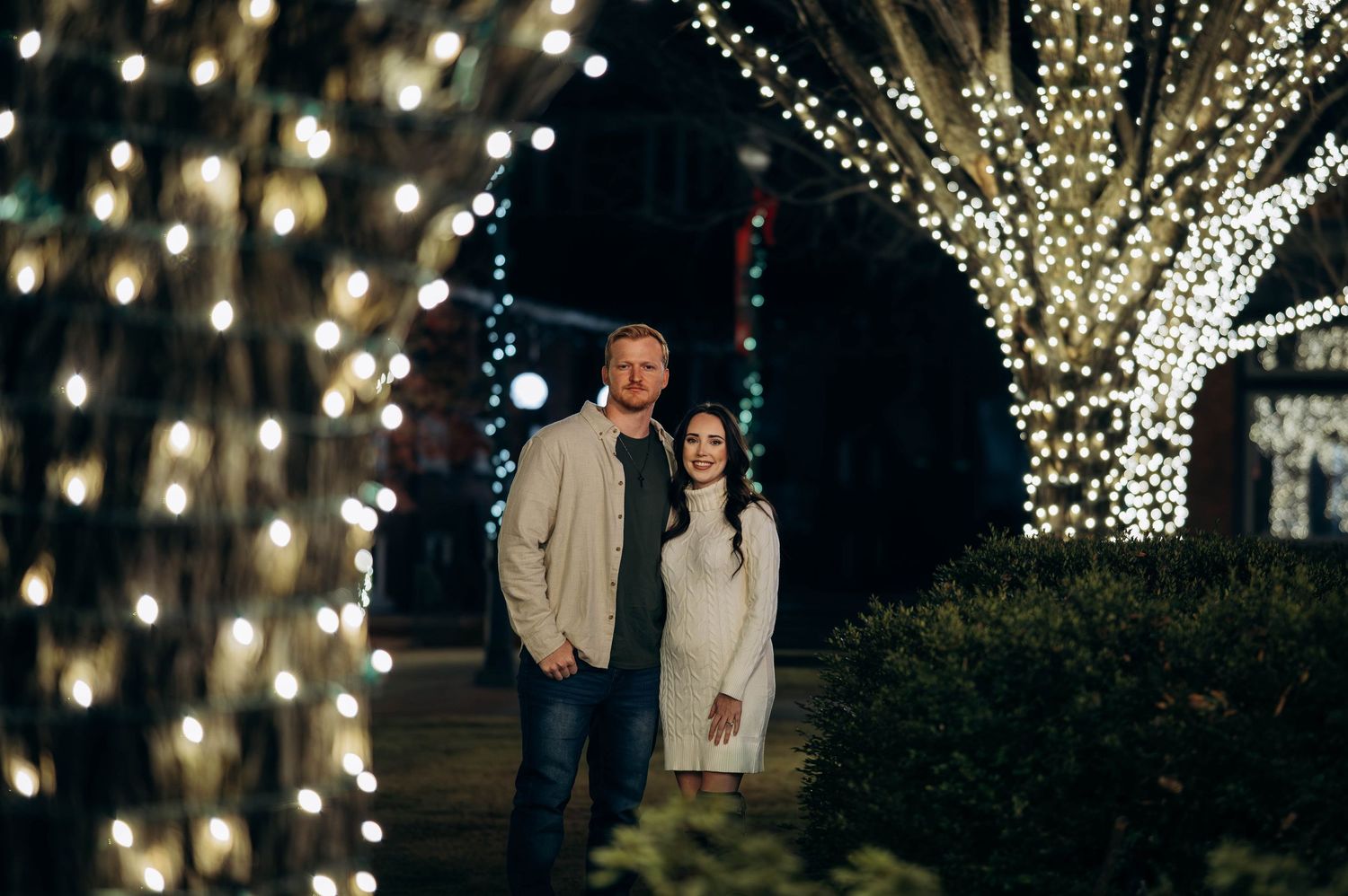 Couple smiling together surrounded by twinkling Christmas lights on trees at night.