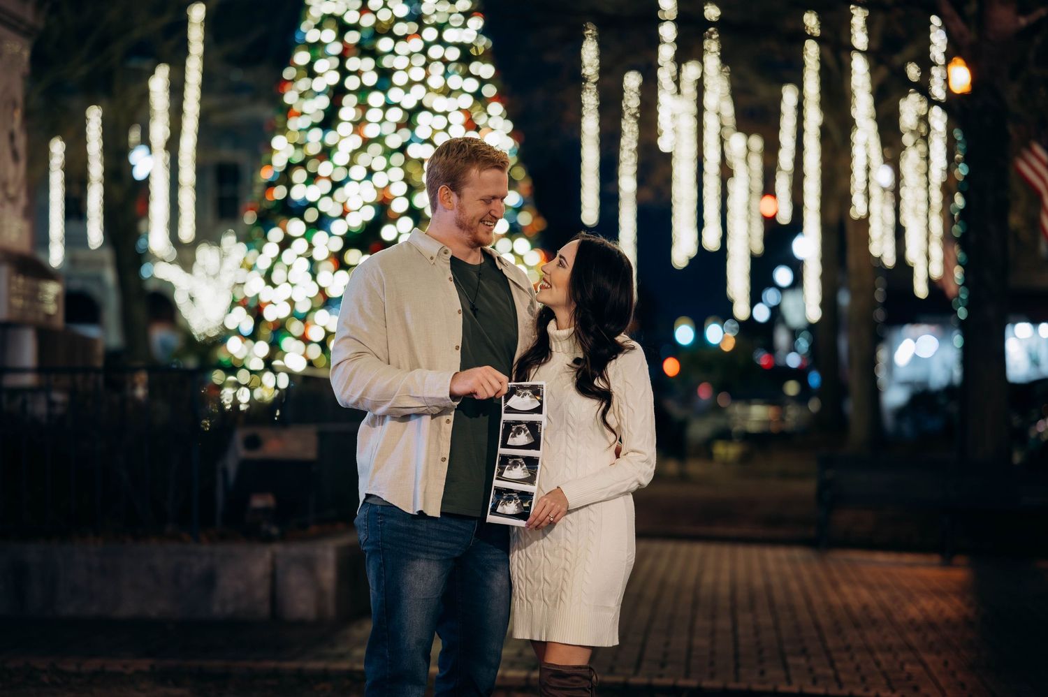 Couple holding ultrasound photos in front of lit Christmas tree at night, announcing pregnancy outdoors.