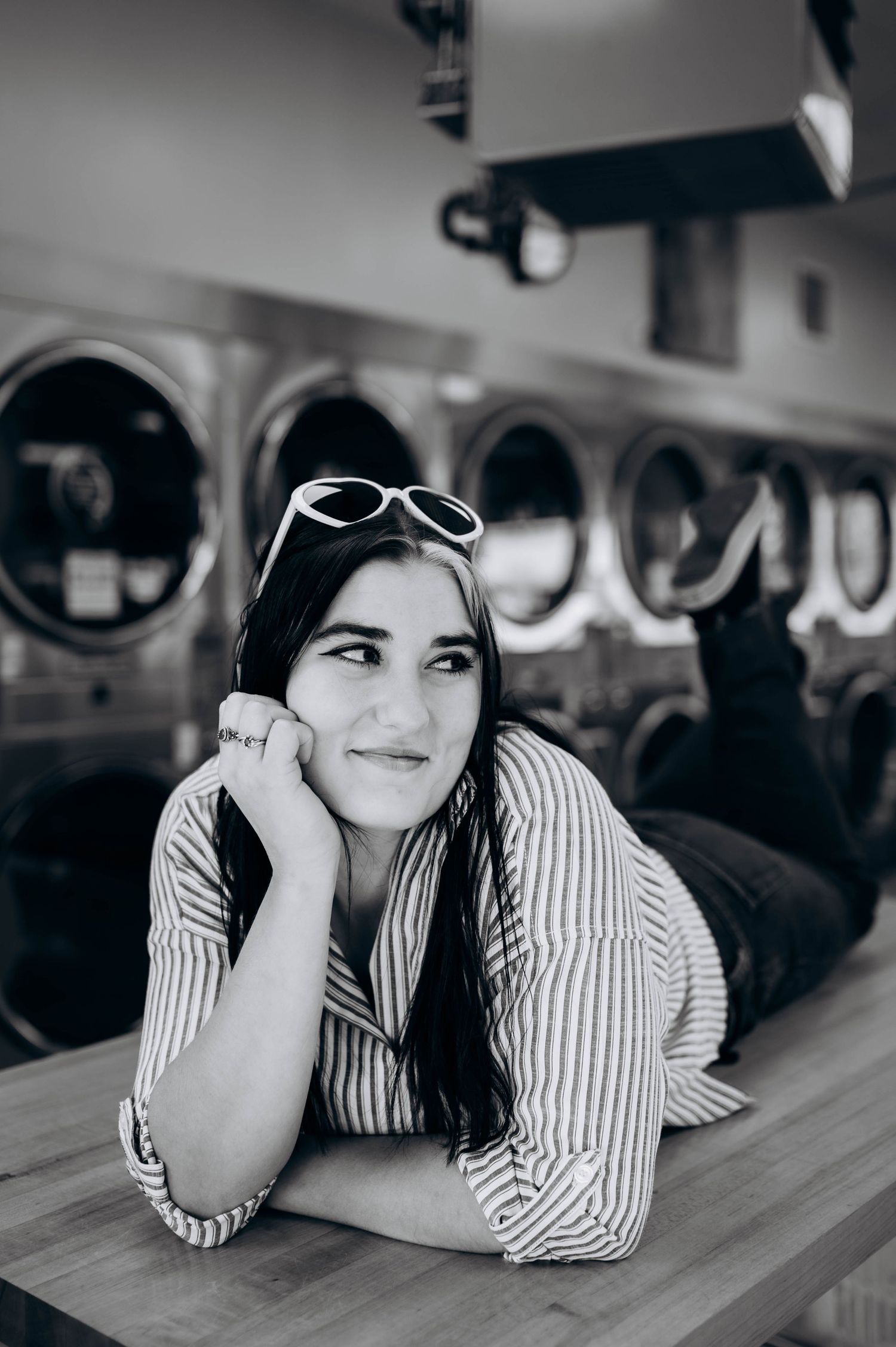 Black and white photo of someone in striped shirt lying on wooden counter at laundromat with vintage washing machines.