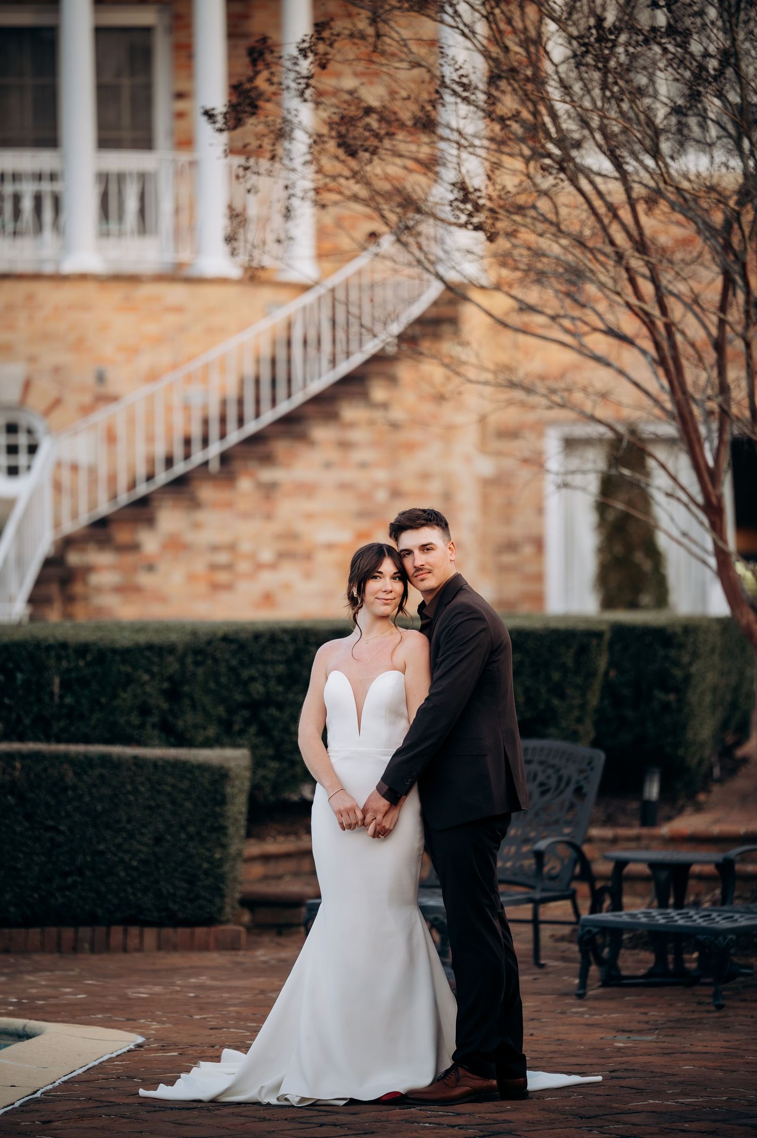 Elegant wedding scene with couple sitting on brick steps near manicured hedges at historic venue.