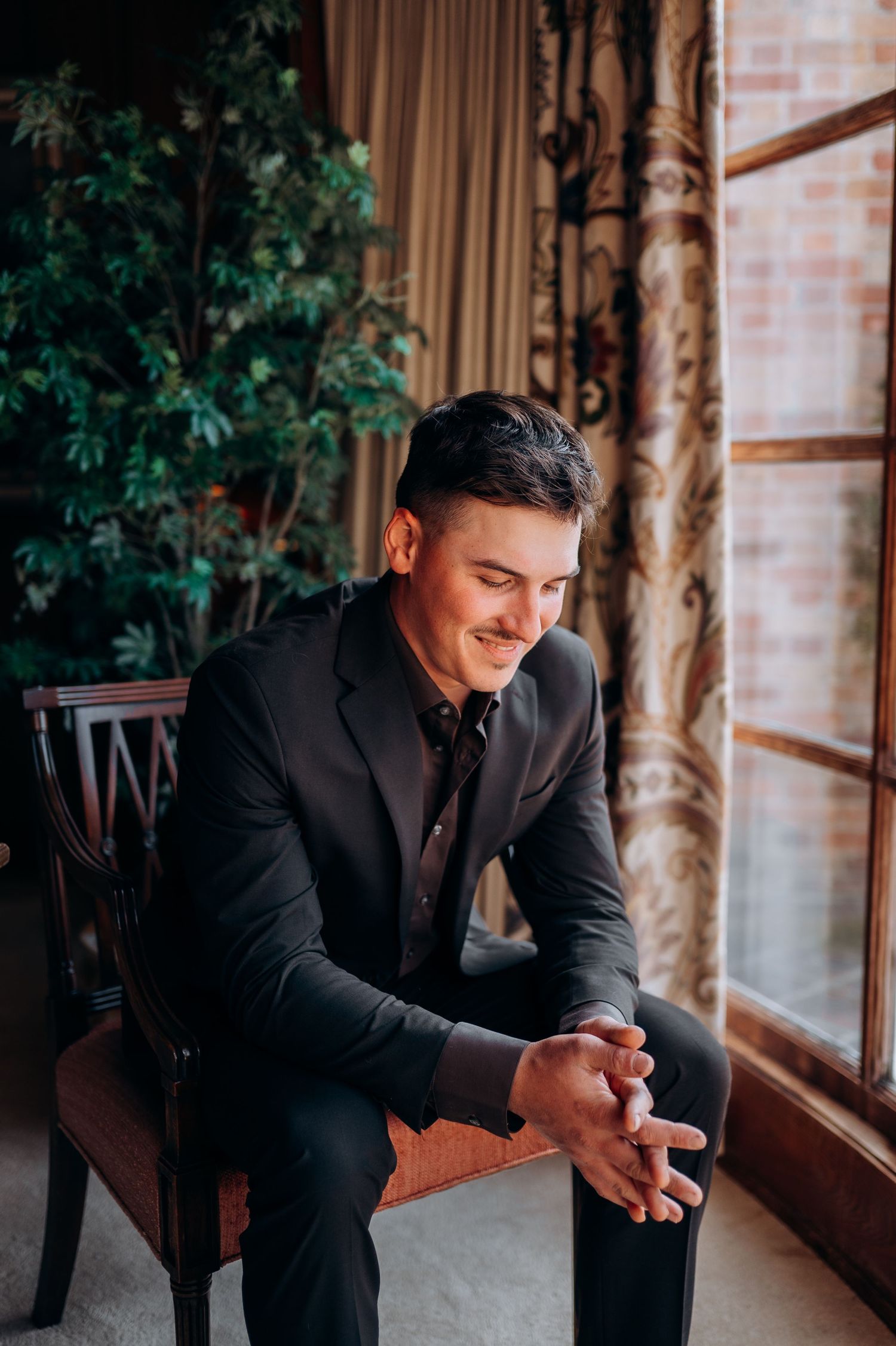 A person in a black suit sits thoughtfully by a window in an elegant interior setting.
