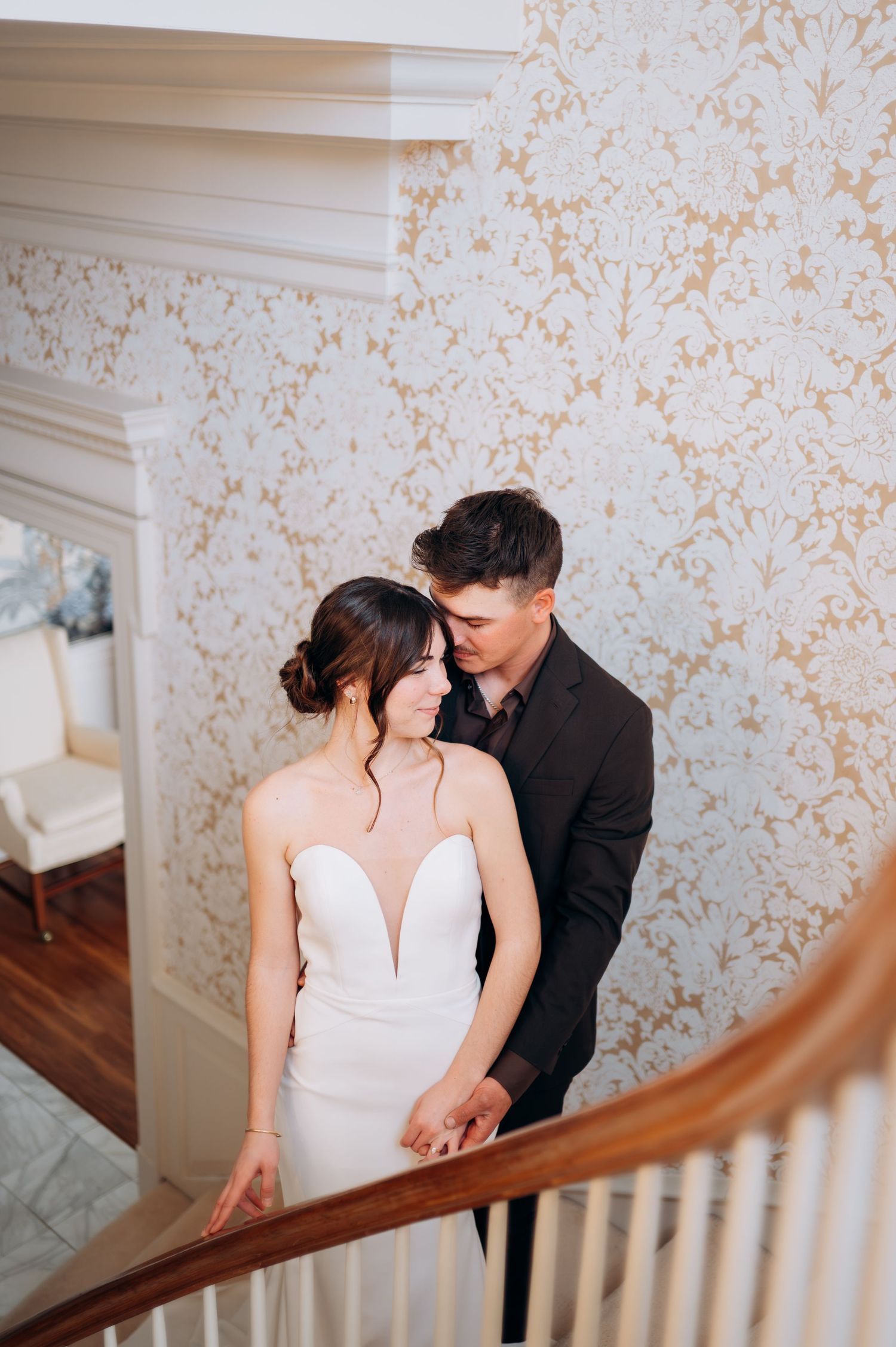 Couple in wedding attire sharing tender moment on staircase with patterned wallpaper.