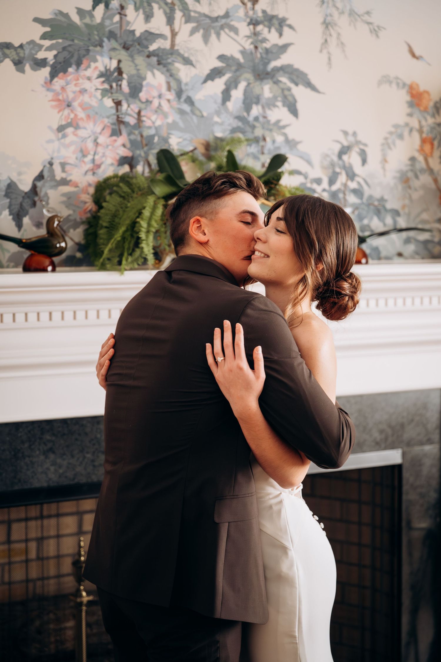 Intimate wedding moment by a fireplace with decorative floral wallpaper.