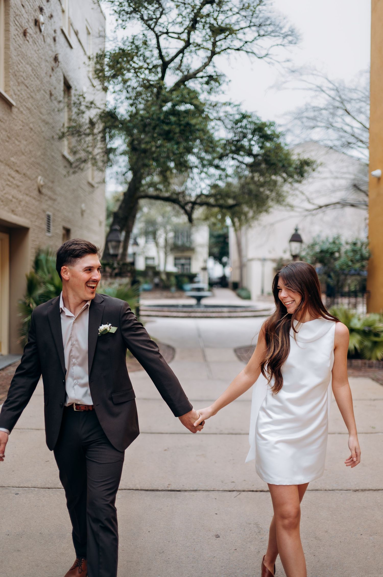 Playful couple in formal attire dancing and laughing together on a paved walkway.