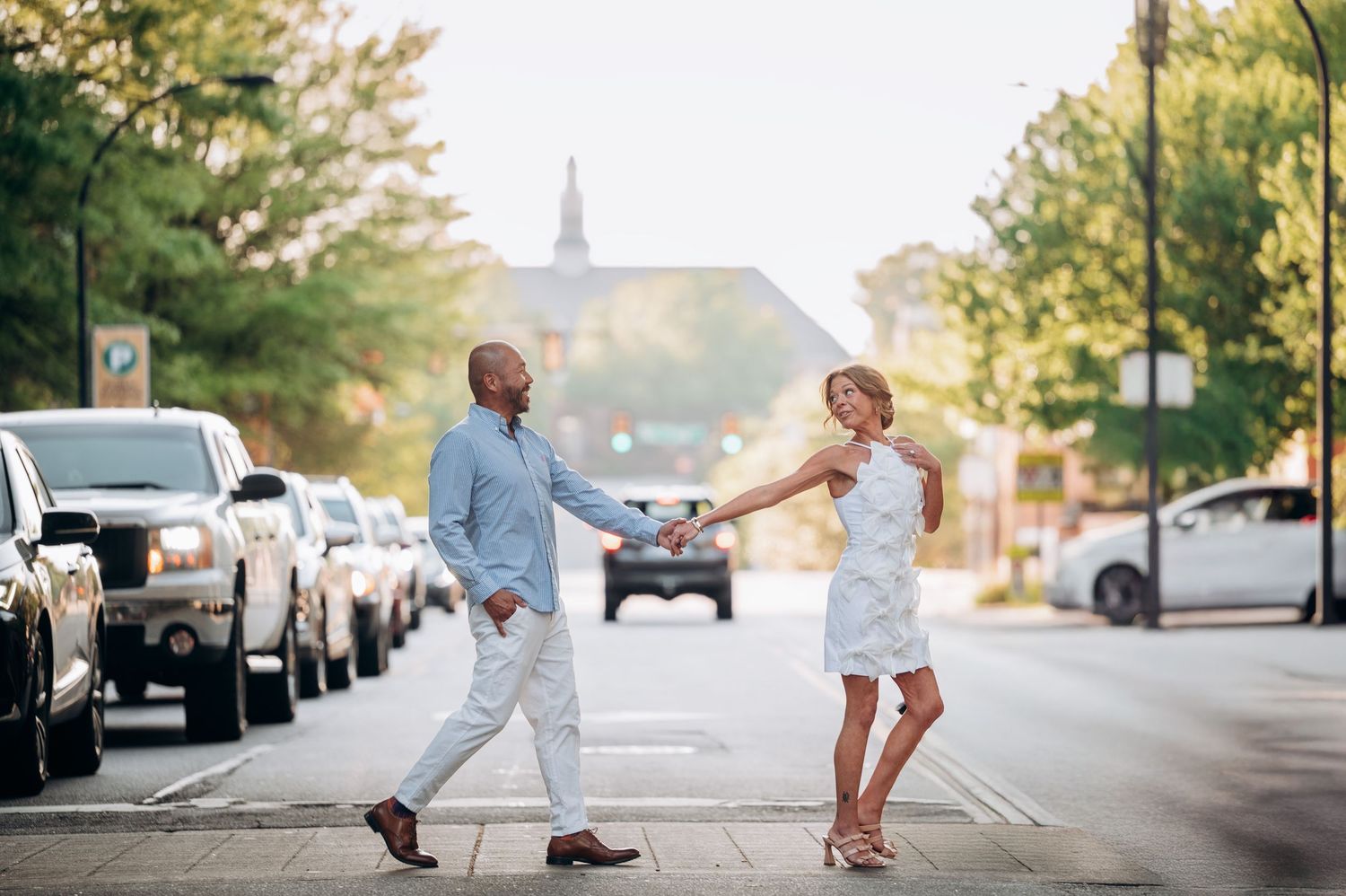 Couple holding hands dancing joyfully on a sunny urban street lined with parked cars.