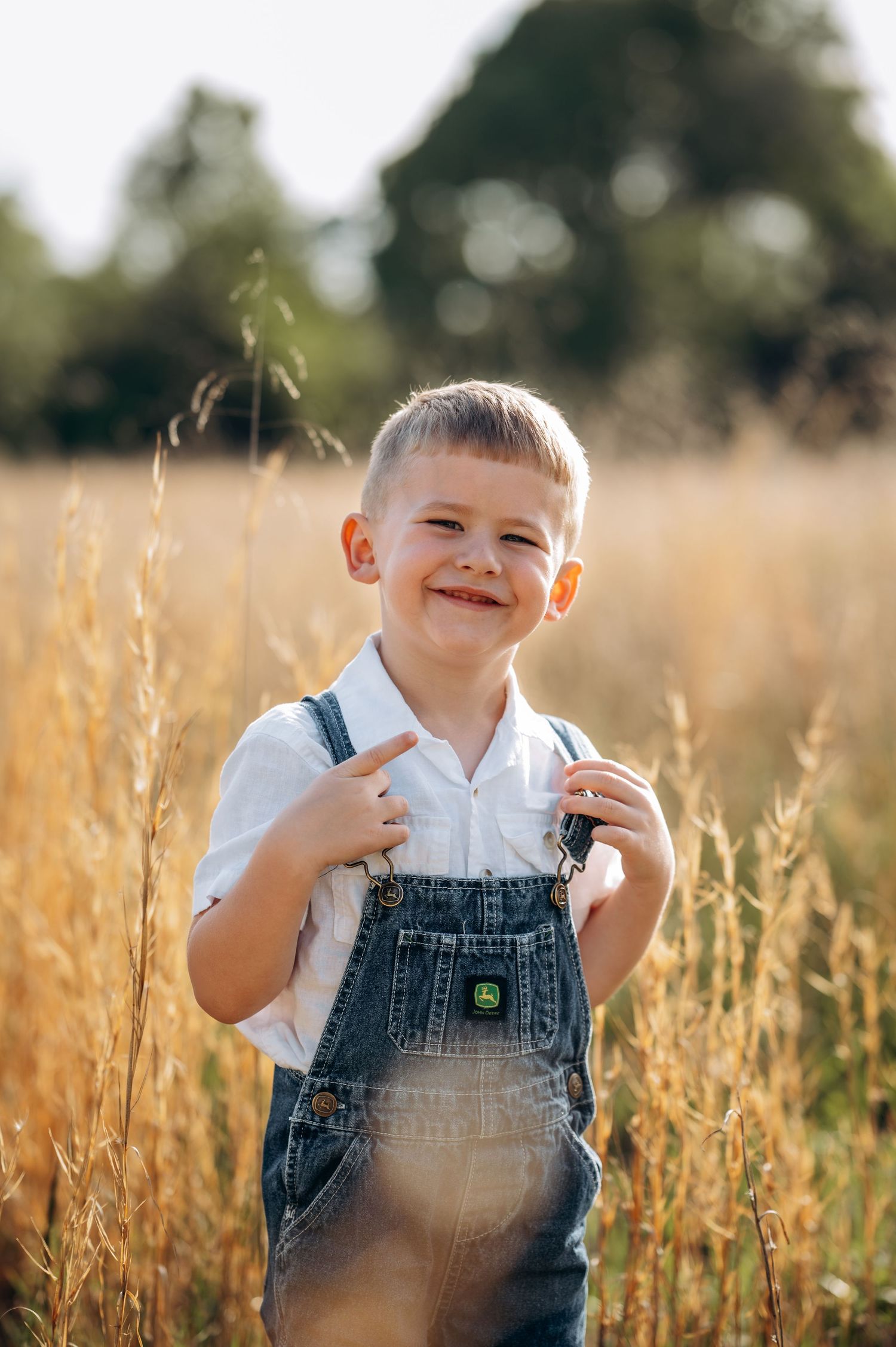 A smiling young boy in denim overalls and white shirt stands in a golden field of tall grass on a sunny day.
