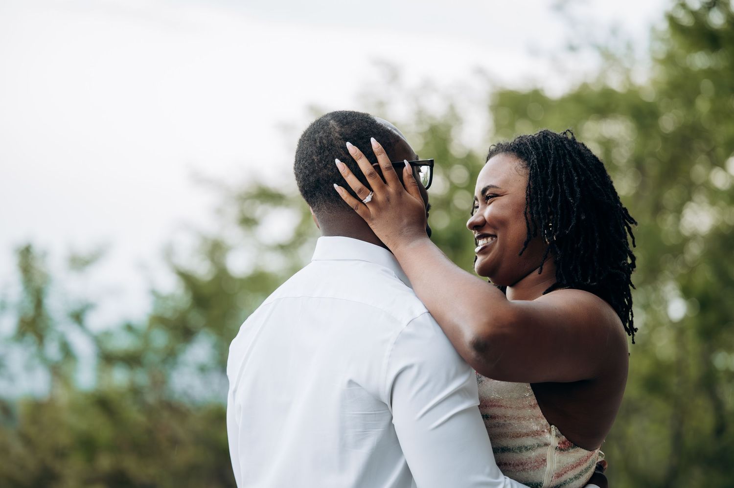A joyful Black couple shares a romantic moment outdoors, woman smiling with engagement ring visible on hand.