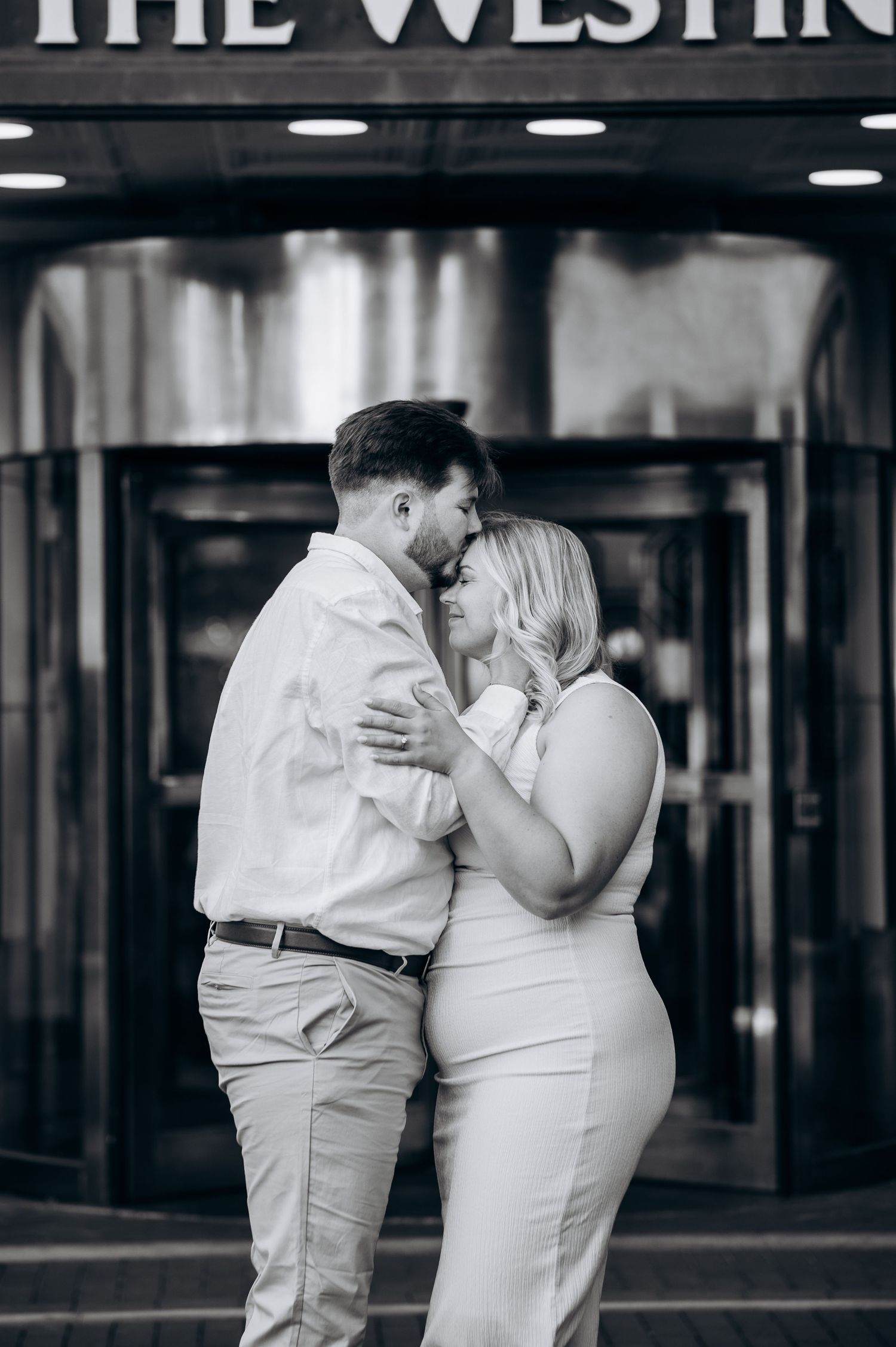 Black and white photo of a couple sharing a tender kiss in front of The Westin hotel entrance.