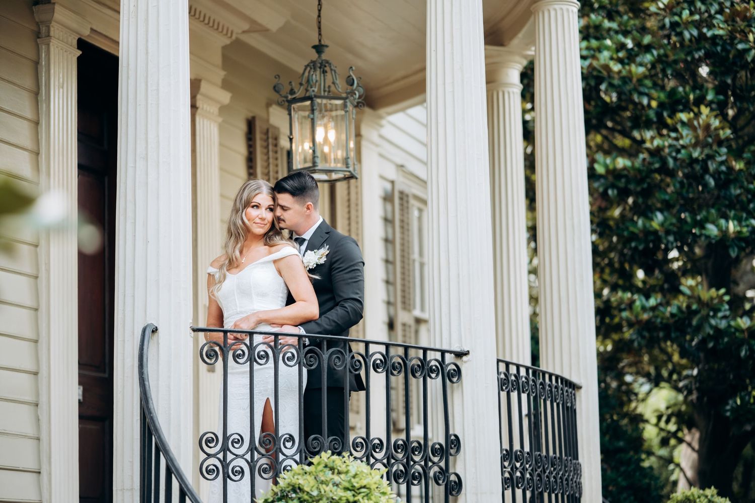 Bride and groom share a romantic moment on an elegant balcony with wrought iron railing and white columns.