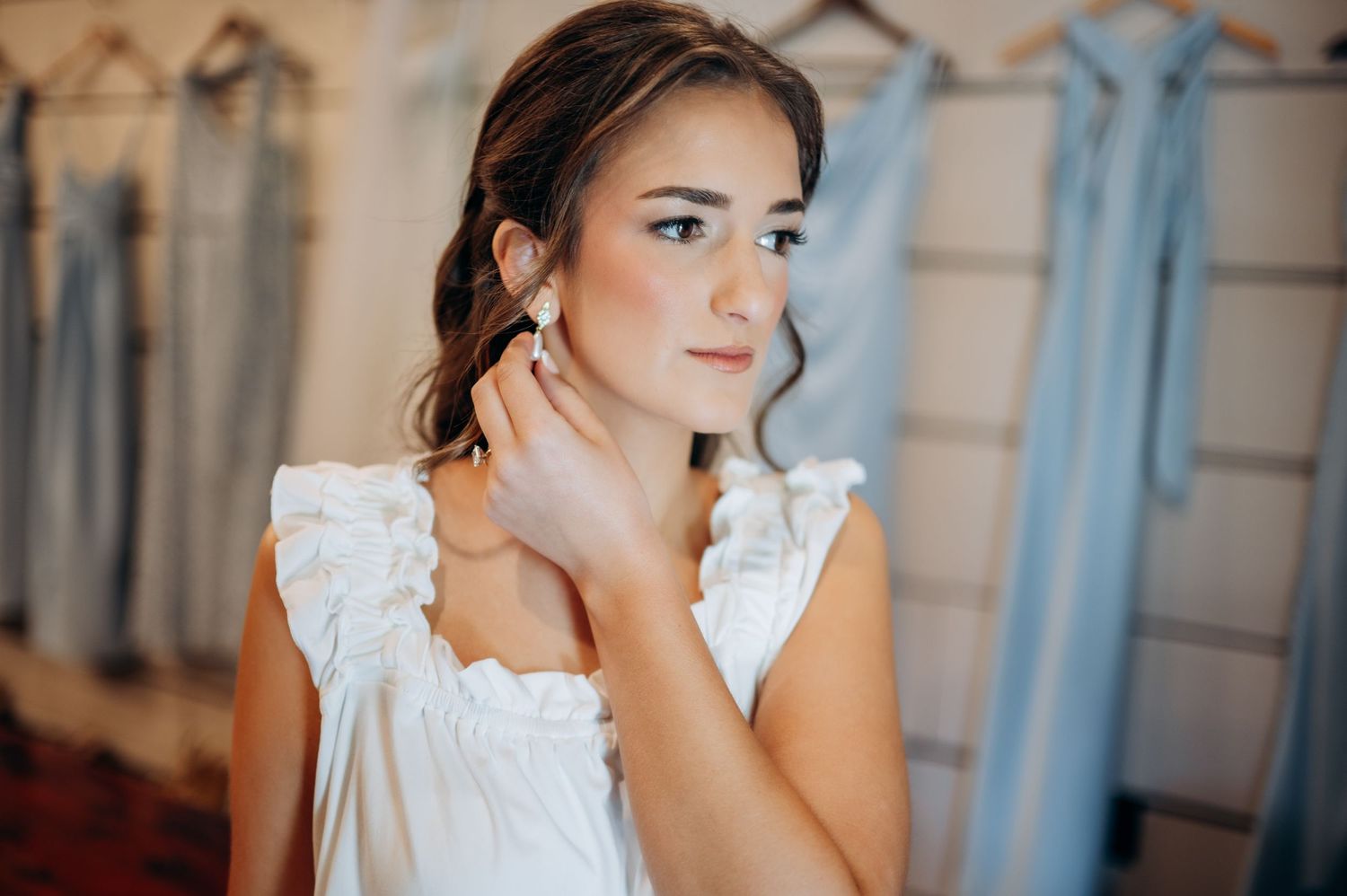 Bride trying on pearl earrings in a bridal boutique, wearing a white ruffled top with blue bridesmaid dresses in background.