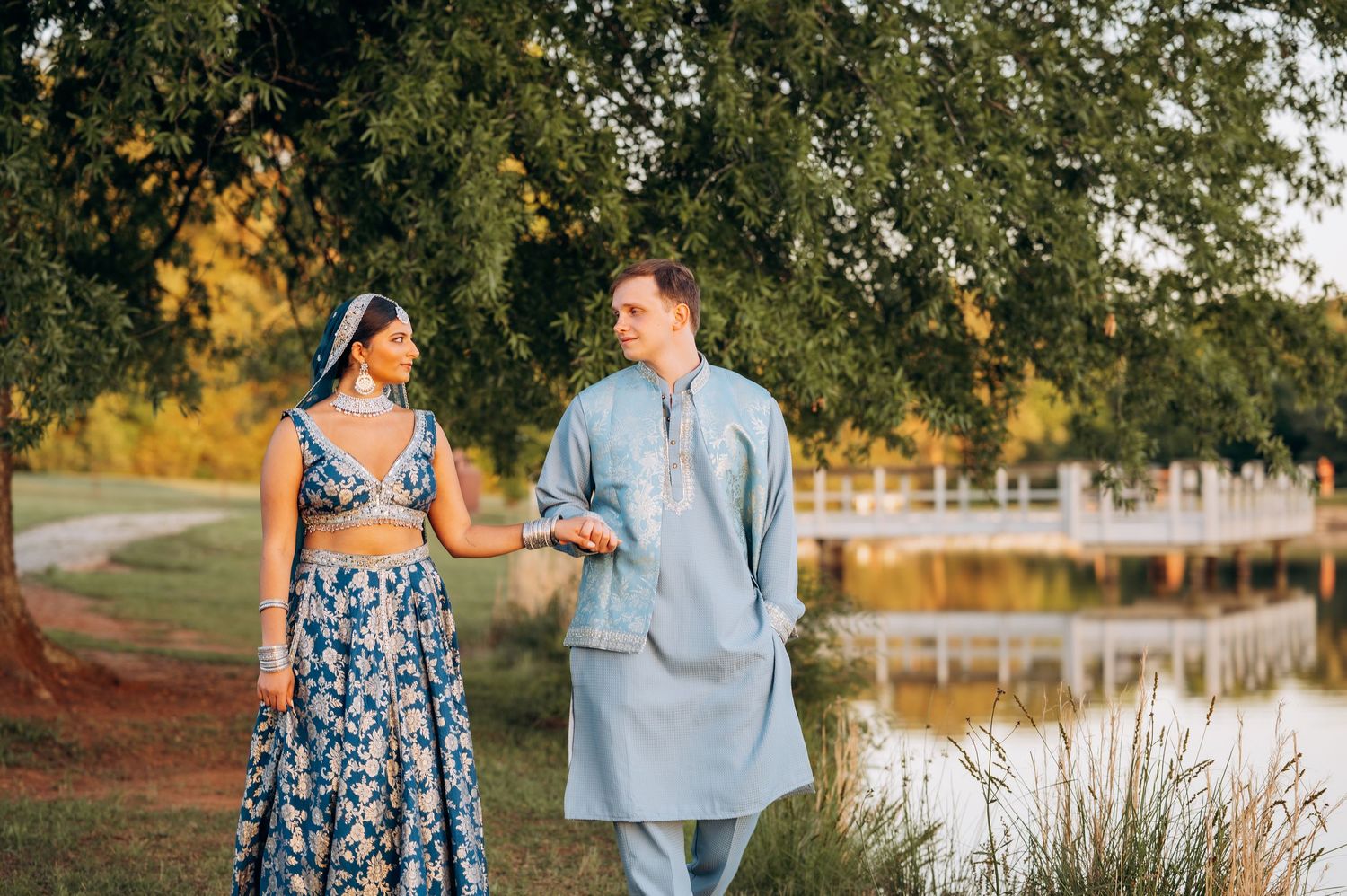 Couple in traditional Indian attire walking hand in hand near a serene lake surrounded by lush green trees.
