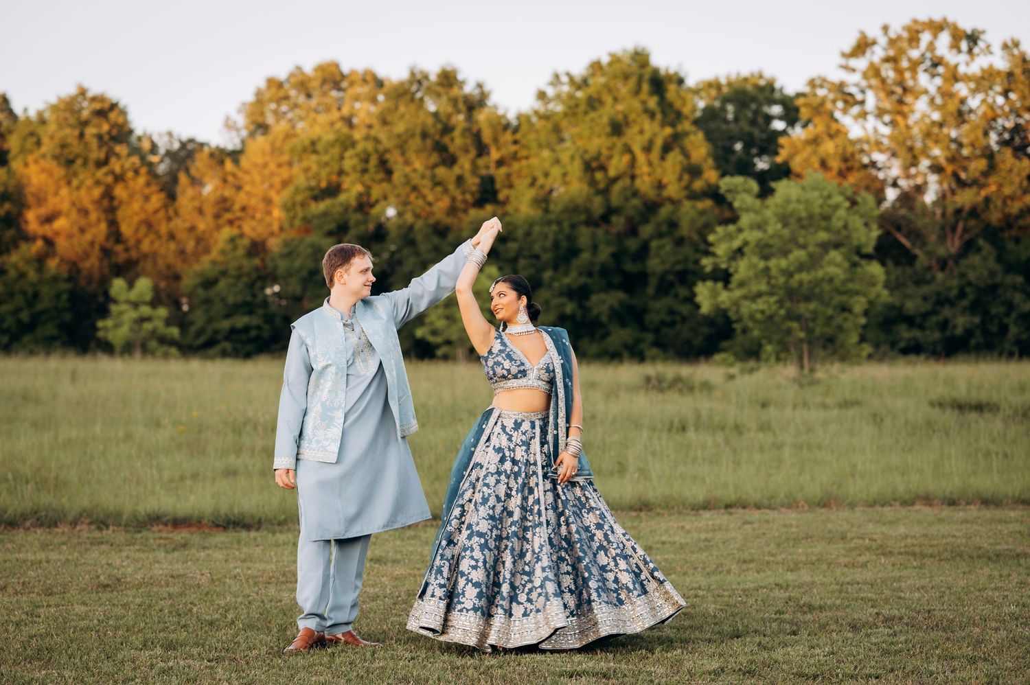 Couple in coordinating blue Indian wedding attire dancing in a green field with colorful autumn trees in the background.