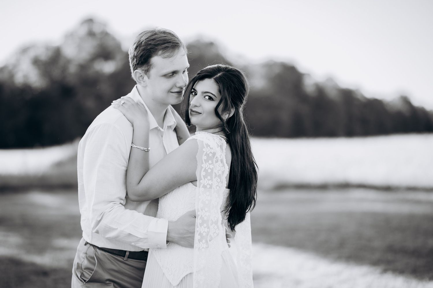 Black and white photo of a couple embracing outdoors, bride in lace dress smiling over shoulder at camera.