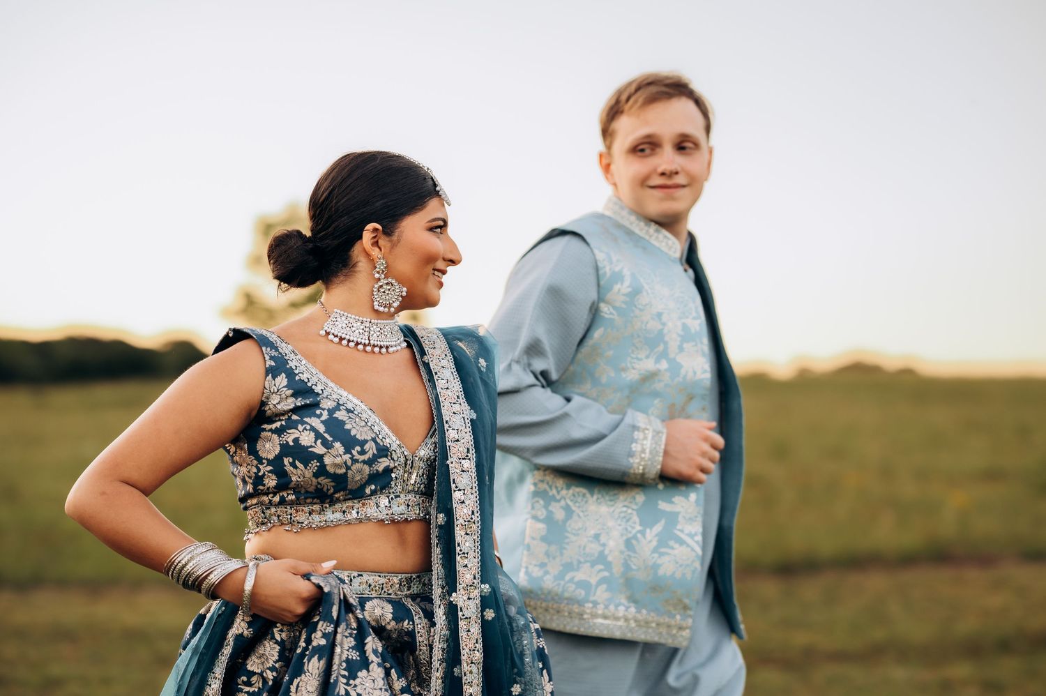 Couple in Indian wedding attire, woman in blue lehenga with jewelry, man in light blue sherwani, posing outdoors at sunset.