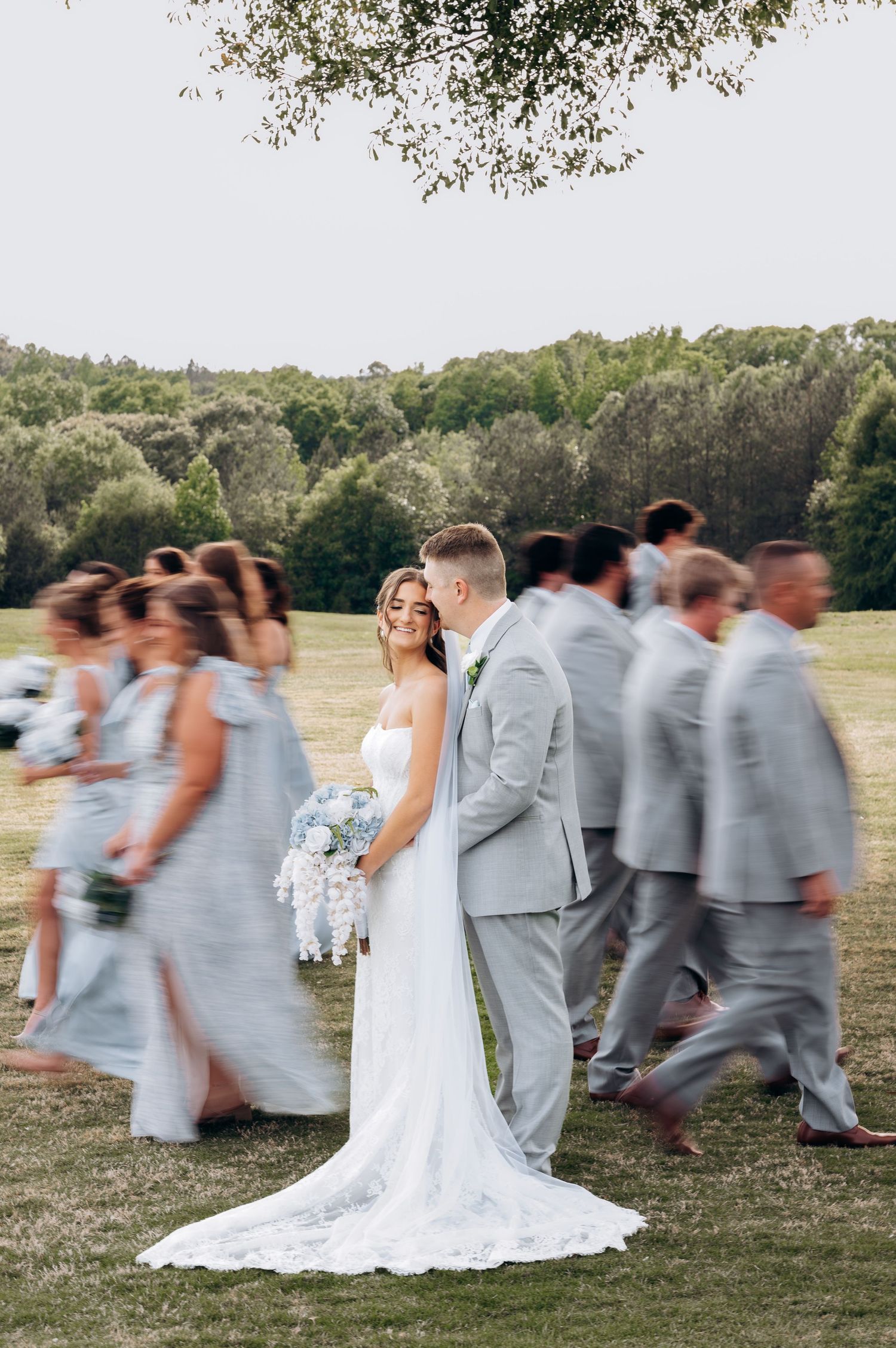 Bride and groom stand still as blurred wedding party walks past on green lawn with trees in background.