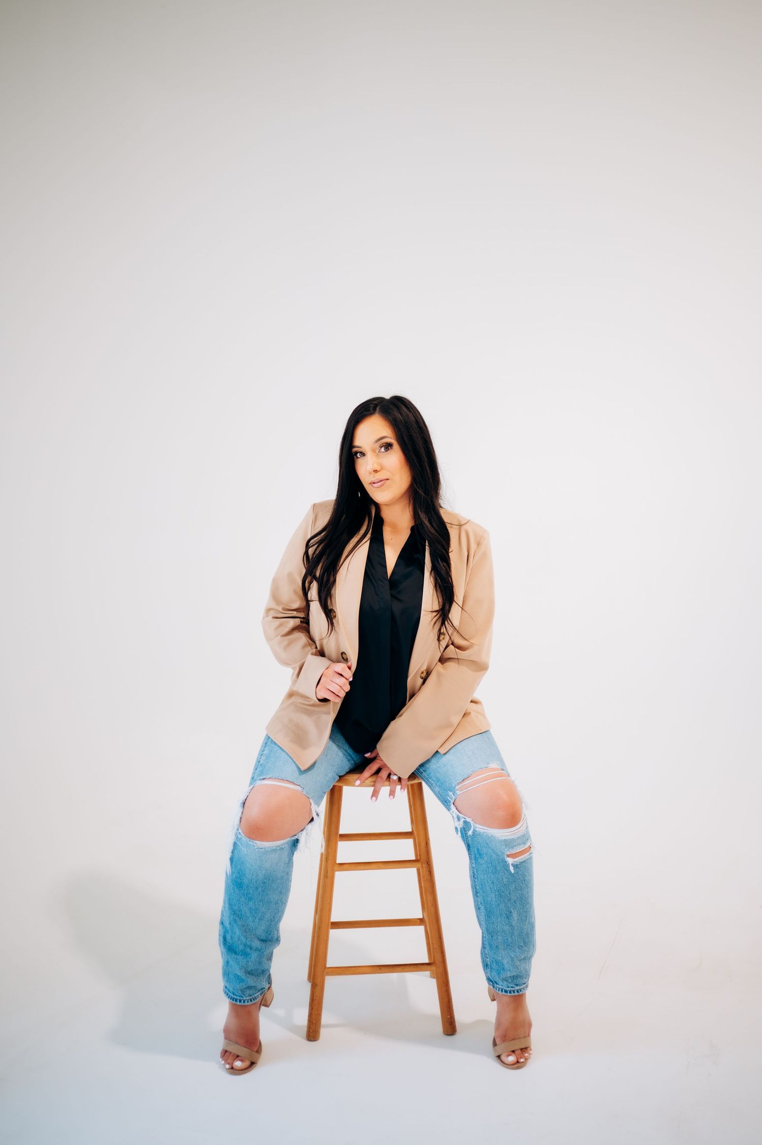 Woman in beige blazer and ripped jeans poses on wooden stool against white background.