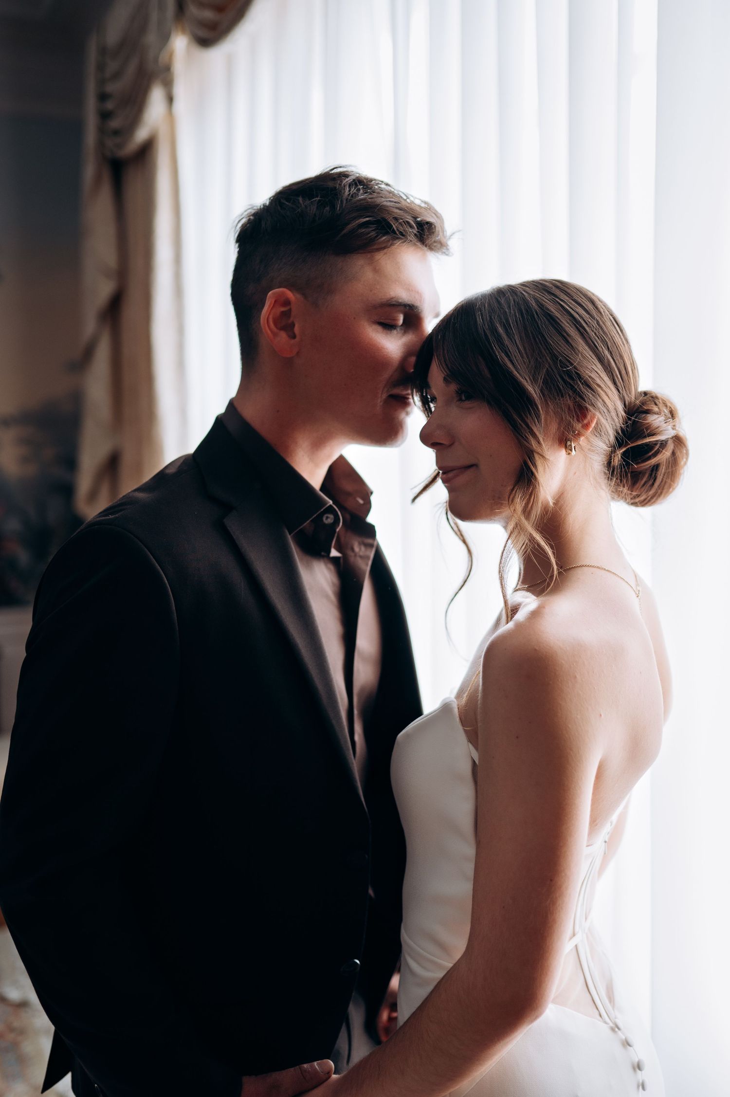 Bride and groom share a tender forehead touch, groom in dark suit, bride in sleek white wedding dress indoors.