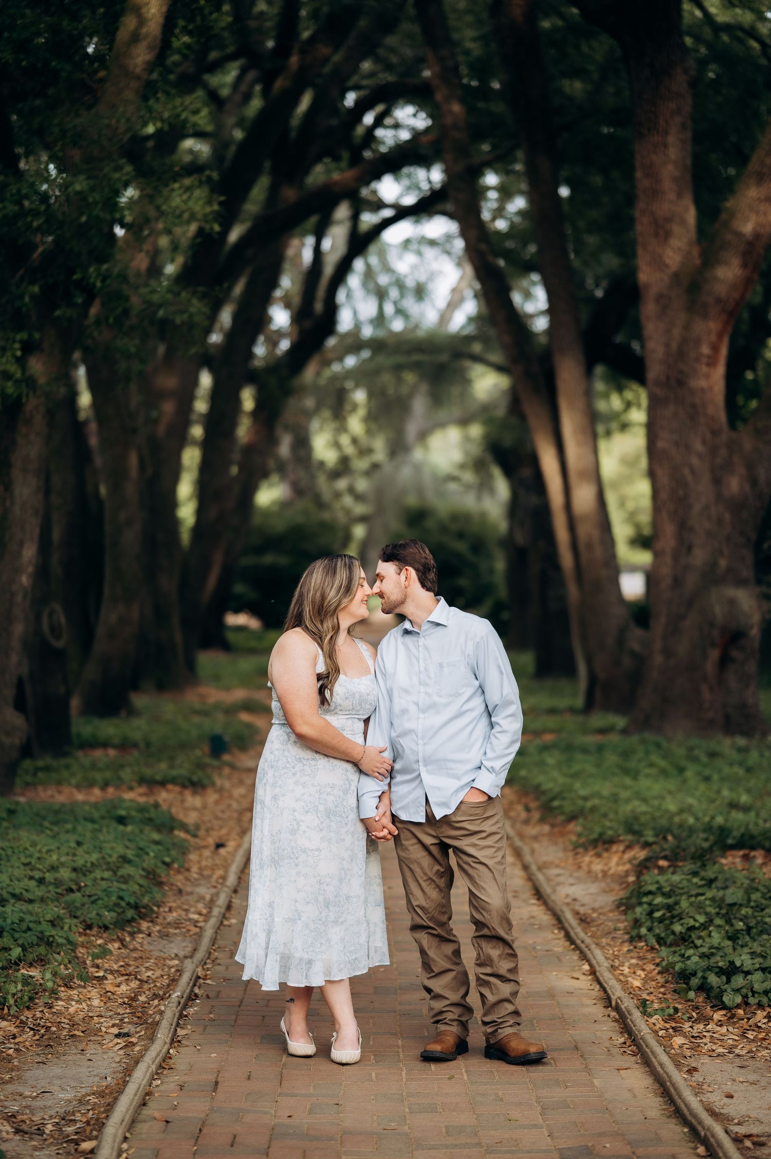Couple sharing a kiss on a brick path surrounded by towering oak trees in a lush garden setting.