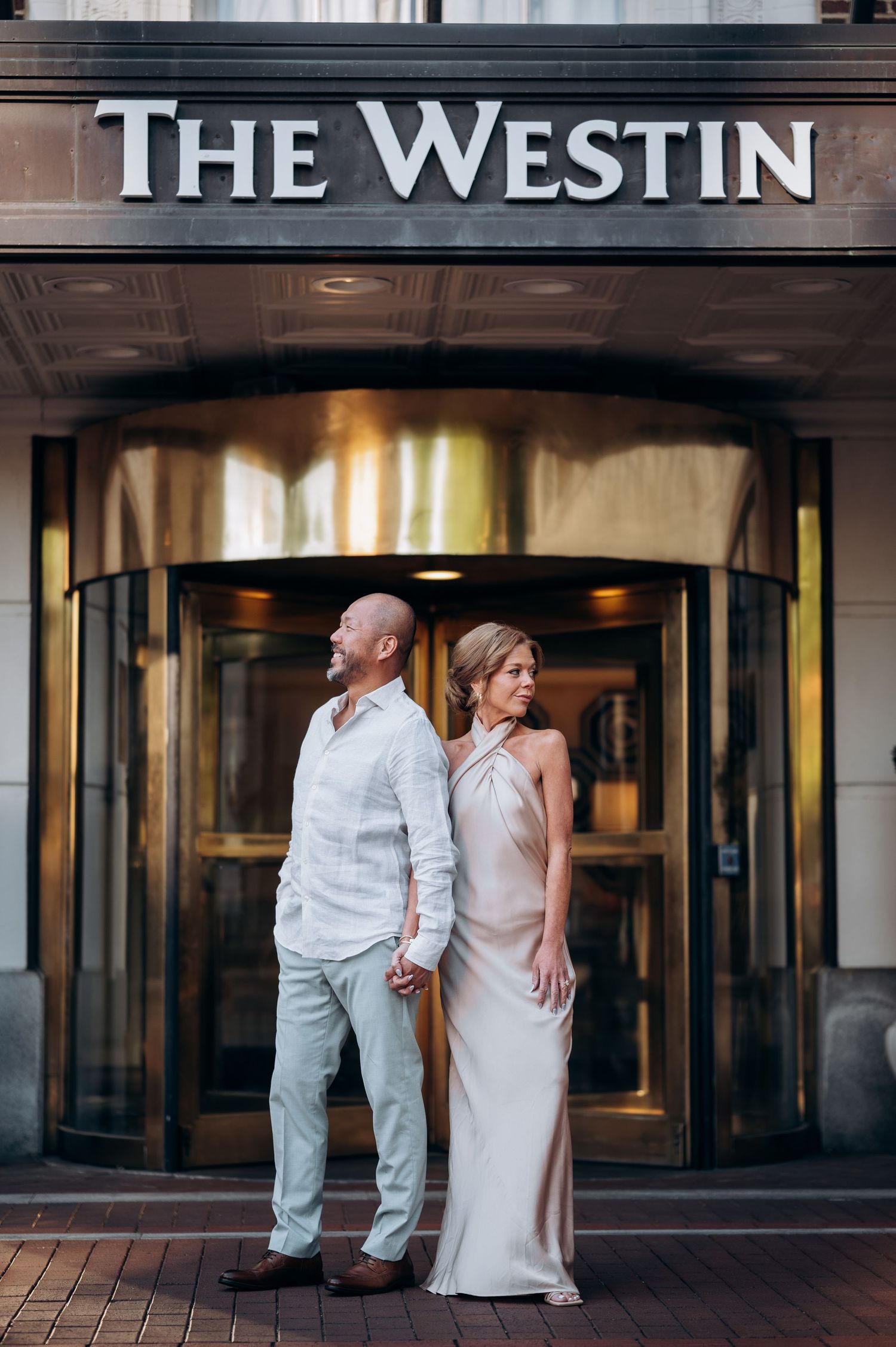 Couple holding hands standing before The Westin hotel entrance, woman in elegant gown and man in linen shirt.