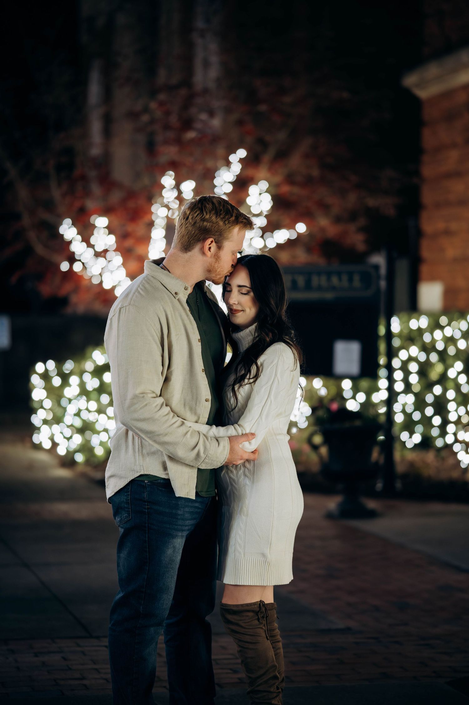 Couple embracing romantically at night with festive holiday lights glowing in the background near City Hall.
