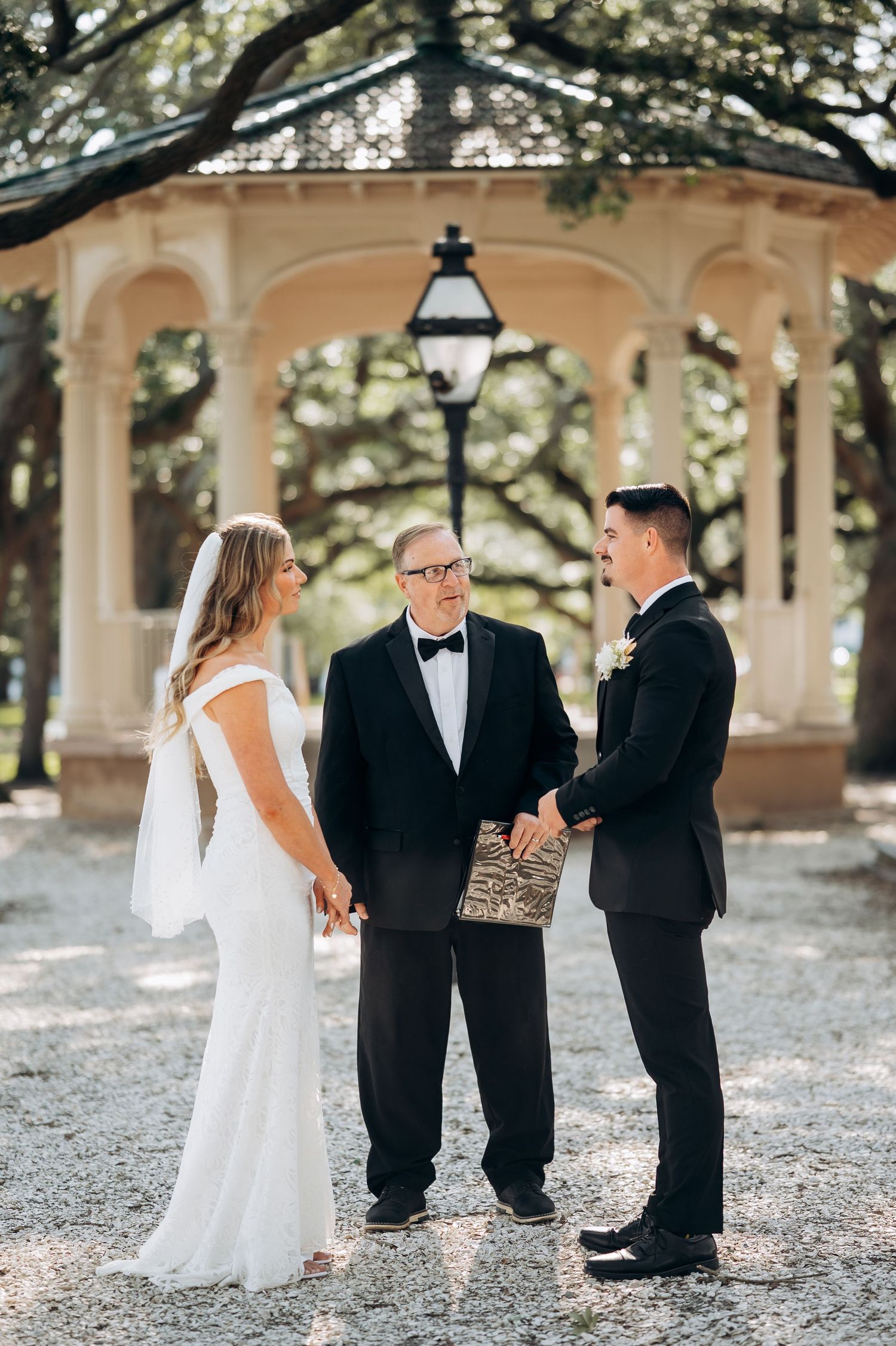 A bride and groom exchange vows with an officiant under a gazebo surrounded by oak trees.