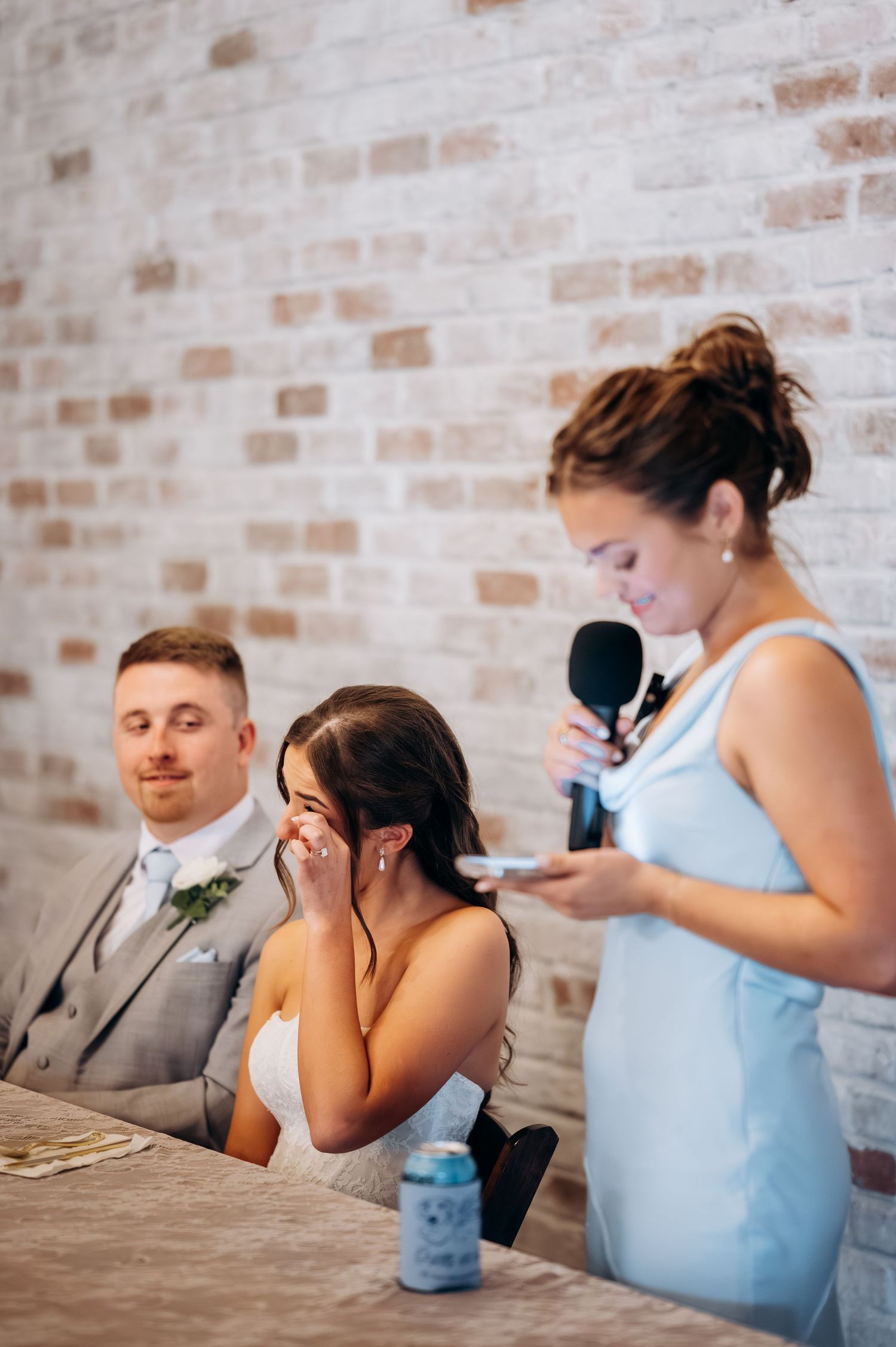 Bride wipes tears during heartfelt maid of honor speech at wedding reception, groom smiling behind her.
