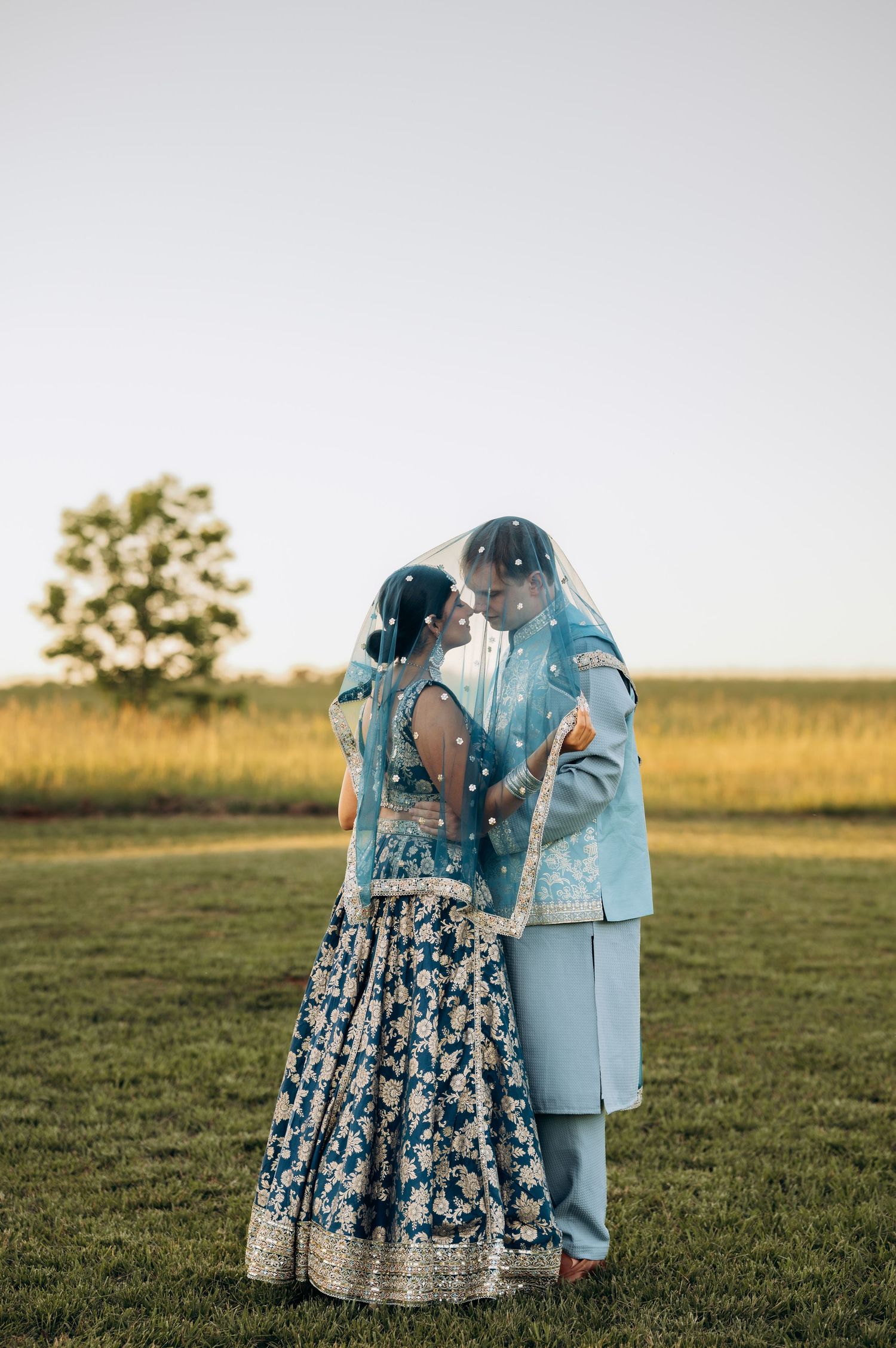 Indian couple in matching blue outfits sharing a kiss under a veil during an outdoor wedding ceremony.