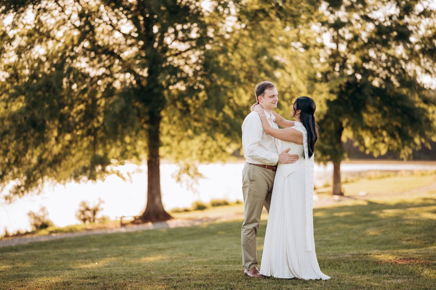Couple embracing in a lush green park near a lake during golden hour in wedding attire.