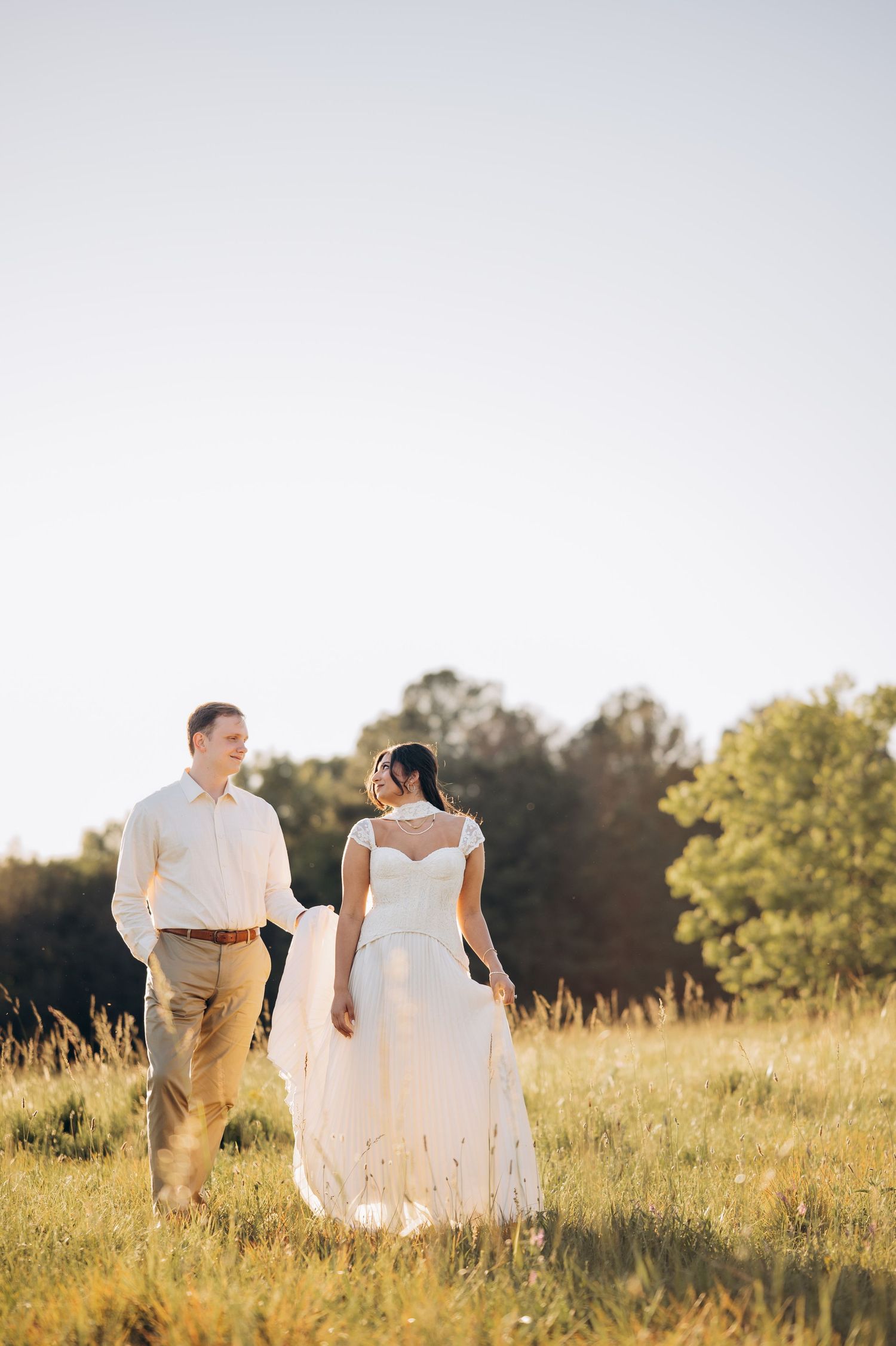 Bride and groom walking hand in hand through a sunlit meadow, bride in white gown, golden hour backdrop.