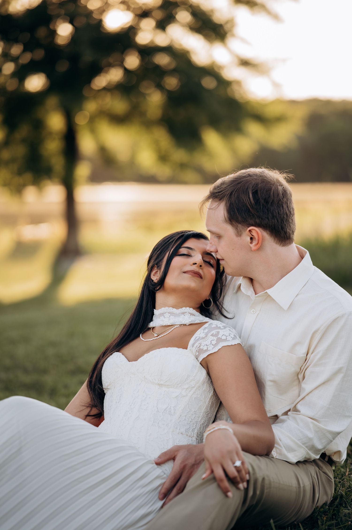 A couple in wedding attire sits together on grass at sunset, the bride leaning back into the groom's embrace.