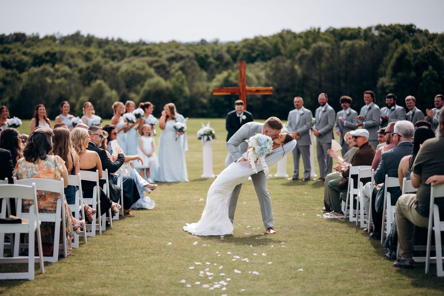 Bride and groom share romantic dip kiss at outdoor wedding ceremony on green lawn with guests seated in white chairs.
