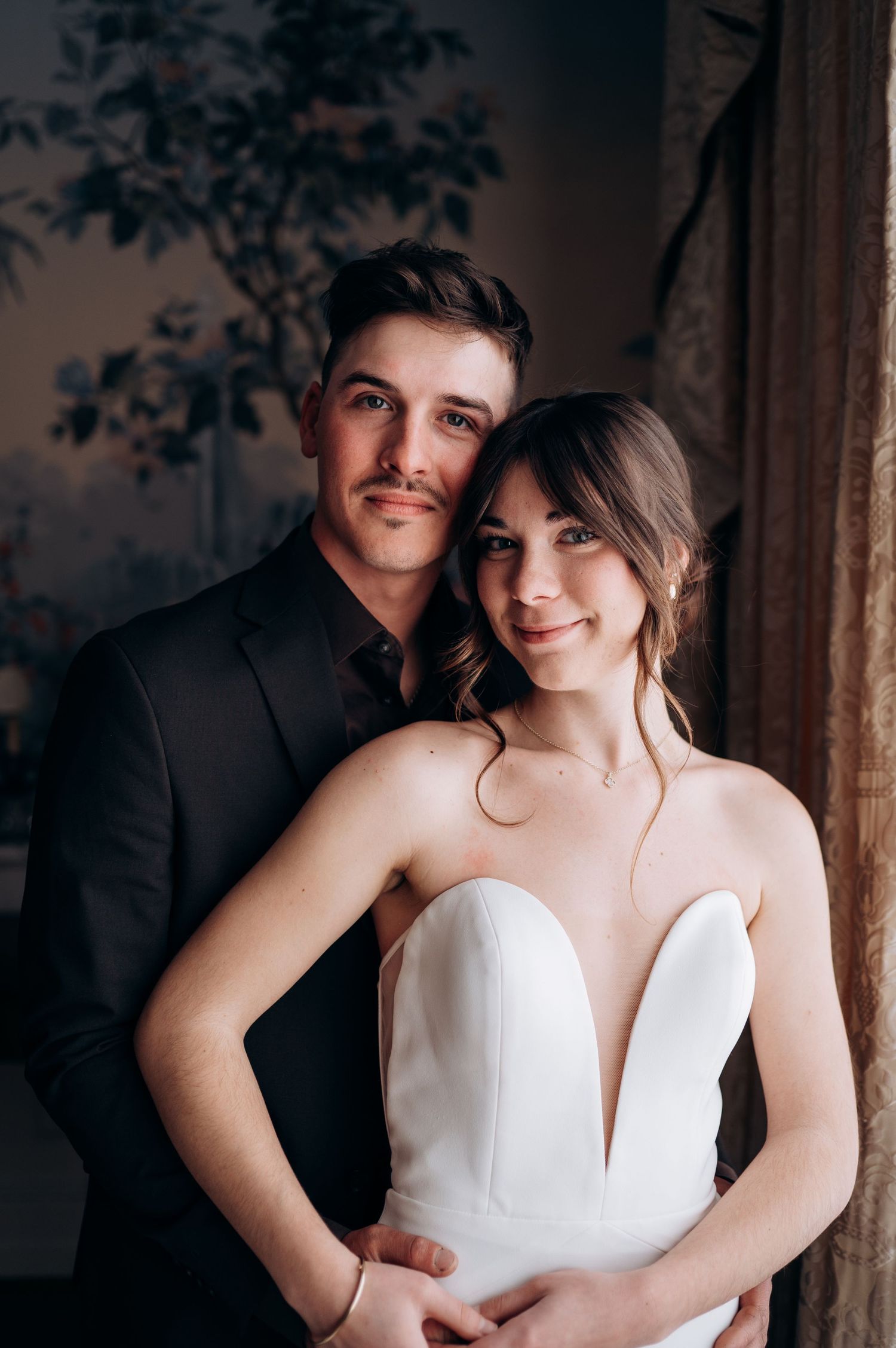 Elegant couple shares romantic moments in formal wedding attire against decorative wallpaper.