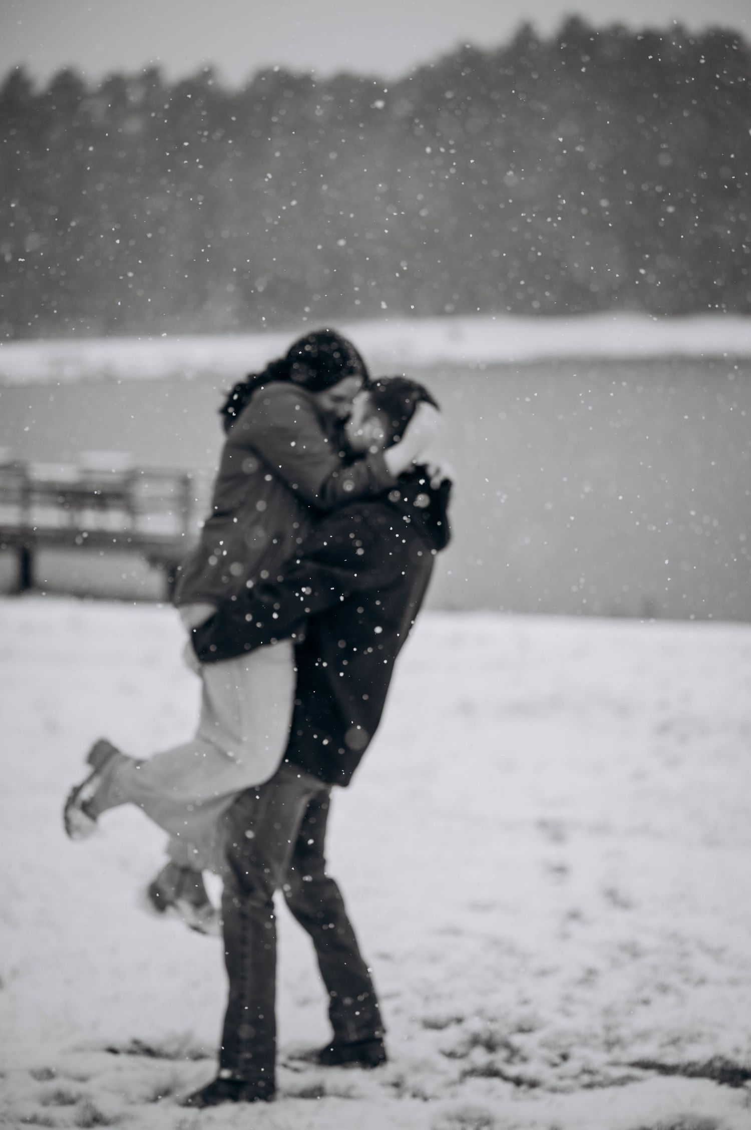 A romantic embrace in the falling snow beside a lake with trees in the distance.