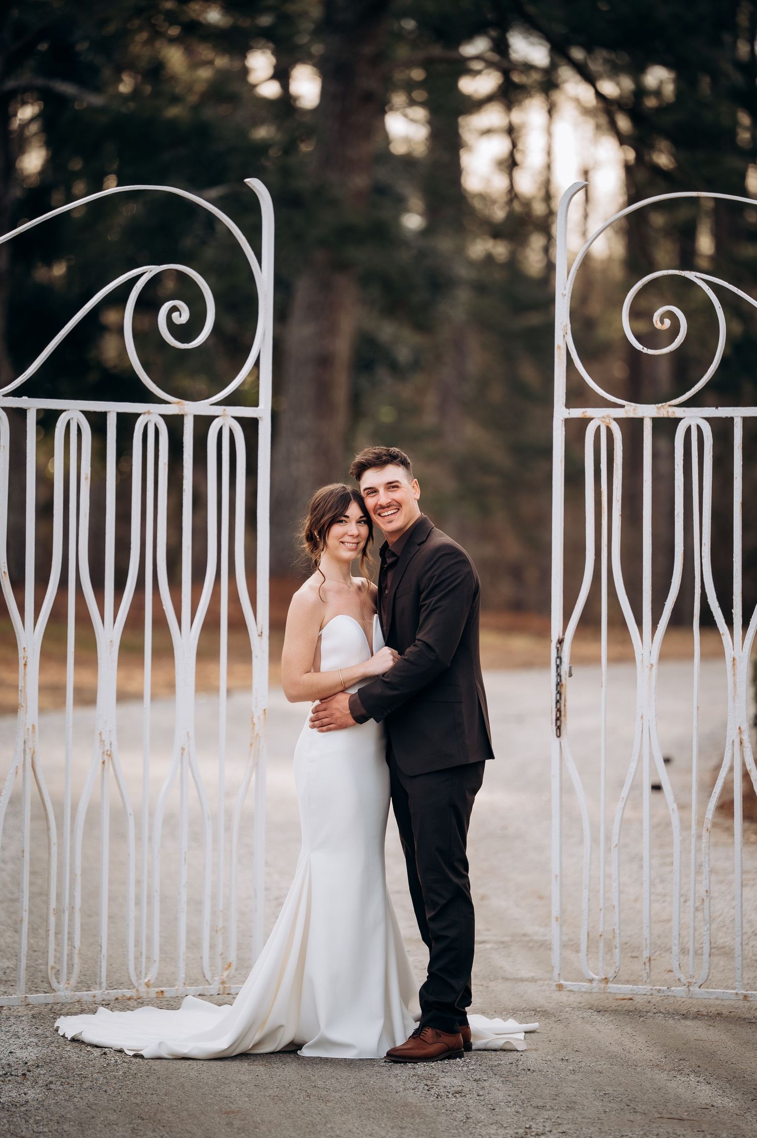 A romantic wedding couple embraces near a decorative white iron fence during sunset.