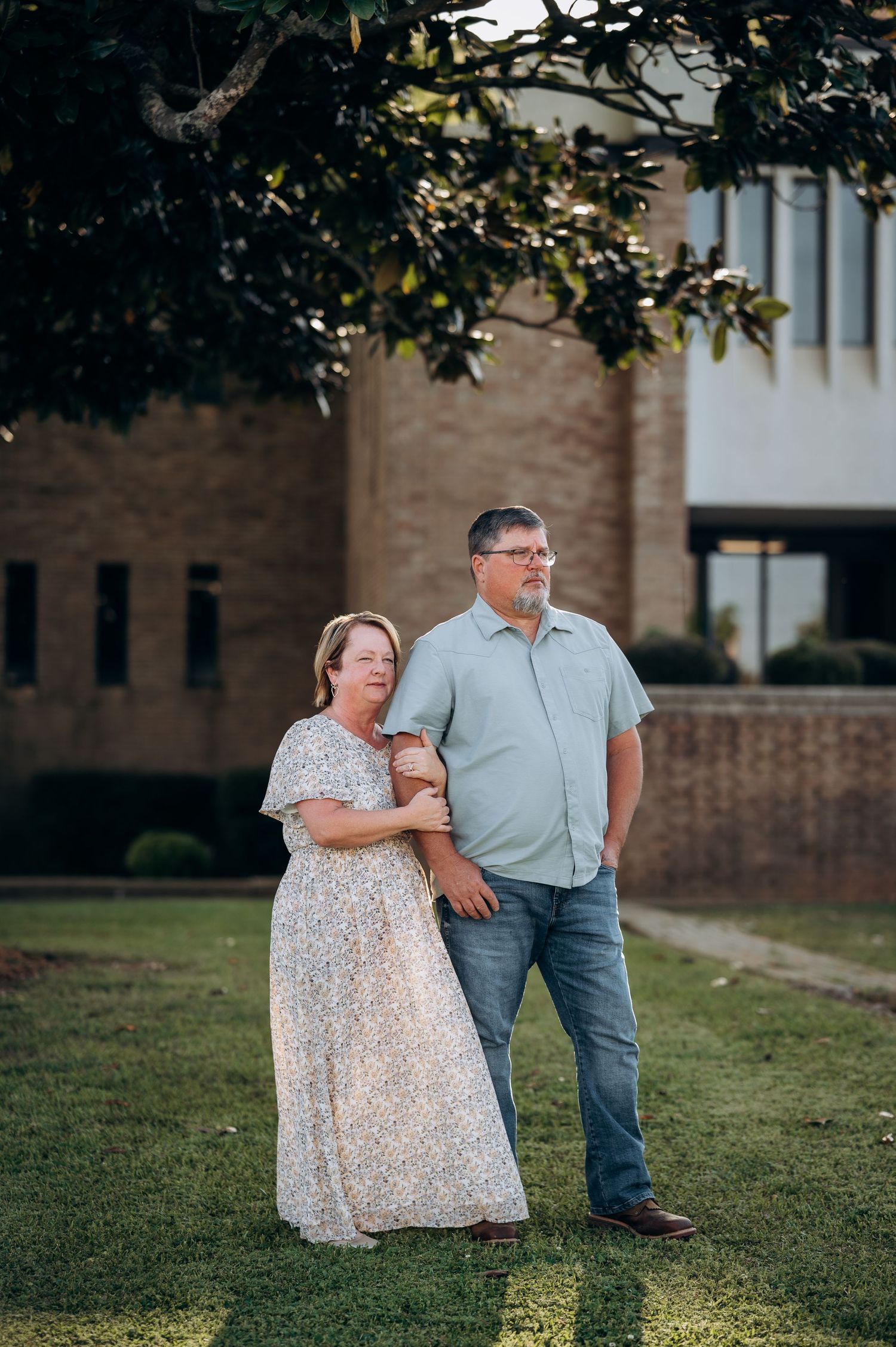 A couple stands together on green grass outside a brick building, woman in floral dress, man in blue shirt and jeans.
