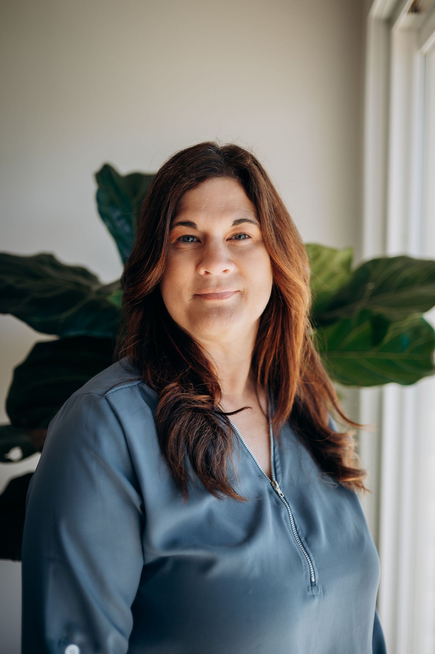 Woman in blue scrubs smiles warmly in front of a large green fiddle leaf fig plant near a bright window.