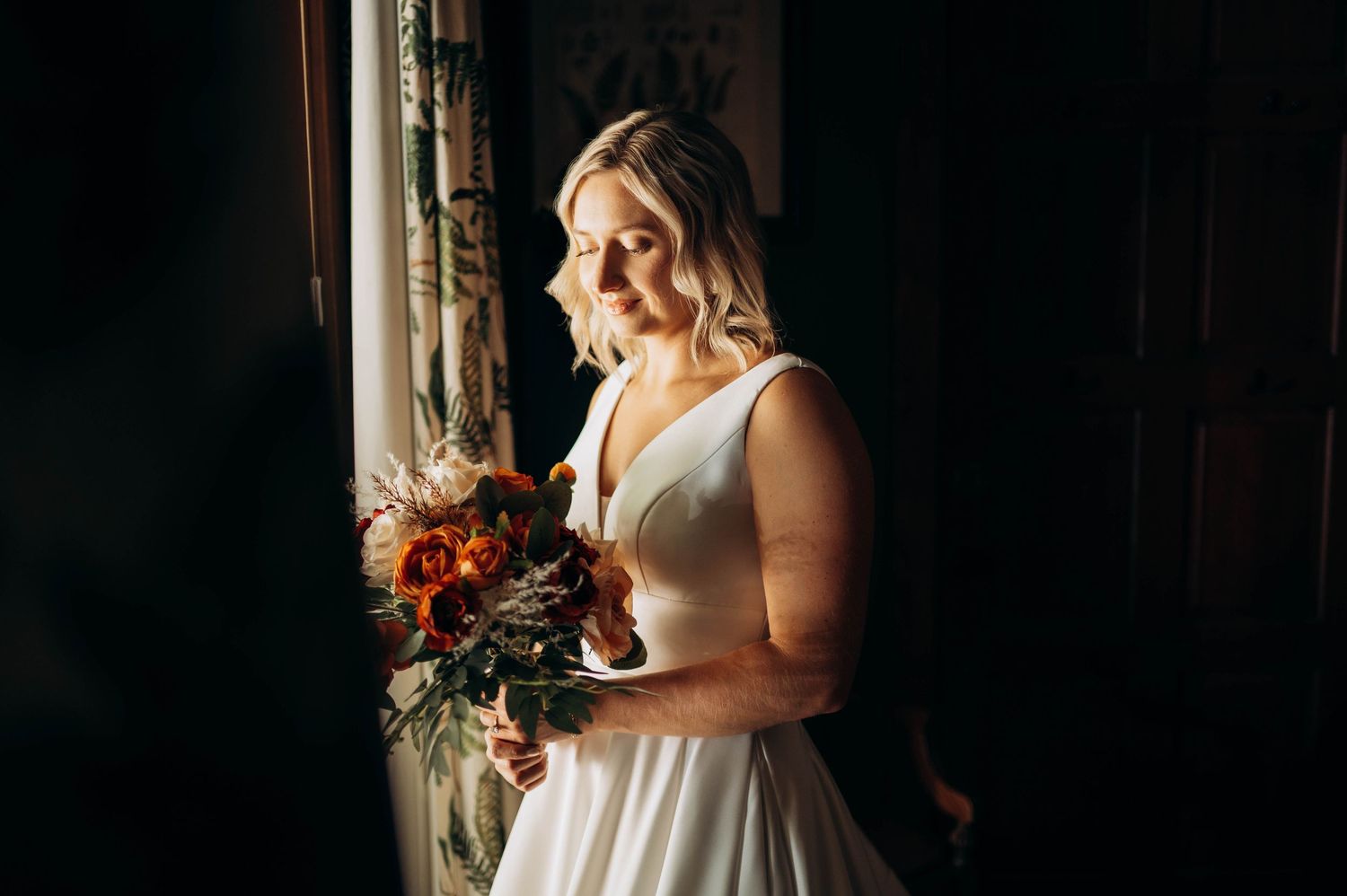 Bride in white dress holds autumn bouquet near window in dark room.