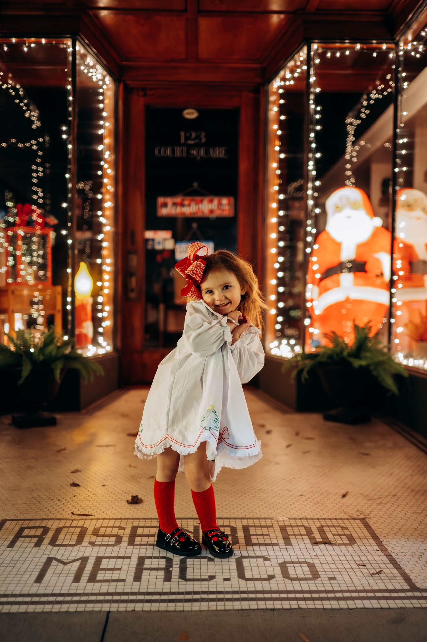 Child in white dress twirling under festive string lights at Rose Creek Mercado.